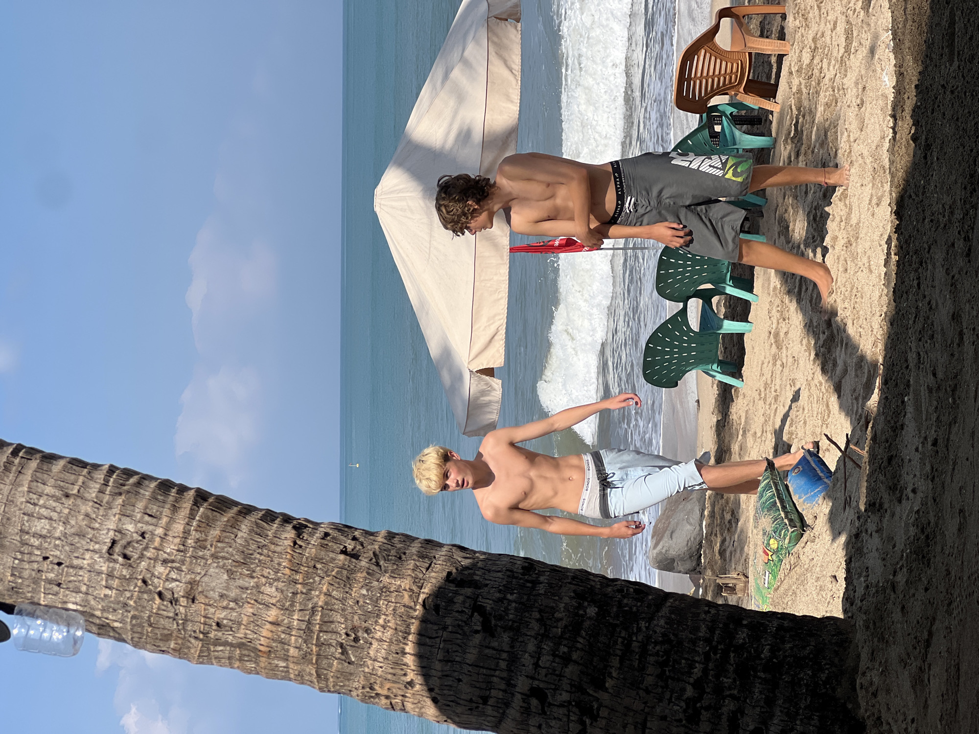 Two young males, appearing to be teenagers, stand on a sandy beach in Kuta, Indonesia. The male on the left has blonde hair and is wearing light blue swimming shorts. The male on the right has brown, curly hair and is wearing dark grey swimming shorts. Both are shirtless. The ocean is visible in the background with waves breaking on the shore. A large beige beach umbrella is situated behind the male on the right. Several green plastic beach chairs and one brown plastic chair are arranged on the sand. A large palm tree trunk is in the foreground on the left, casting a shadow. Other objects on the beach include a blue object, a green mat, and some debris. The sky is blue with scattered clouds.