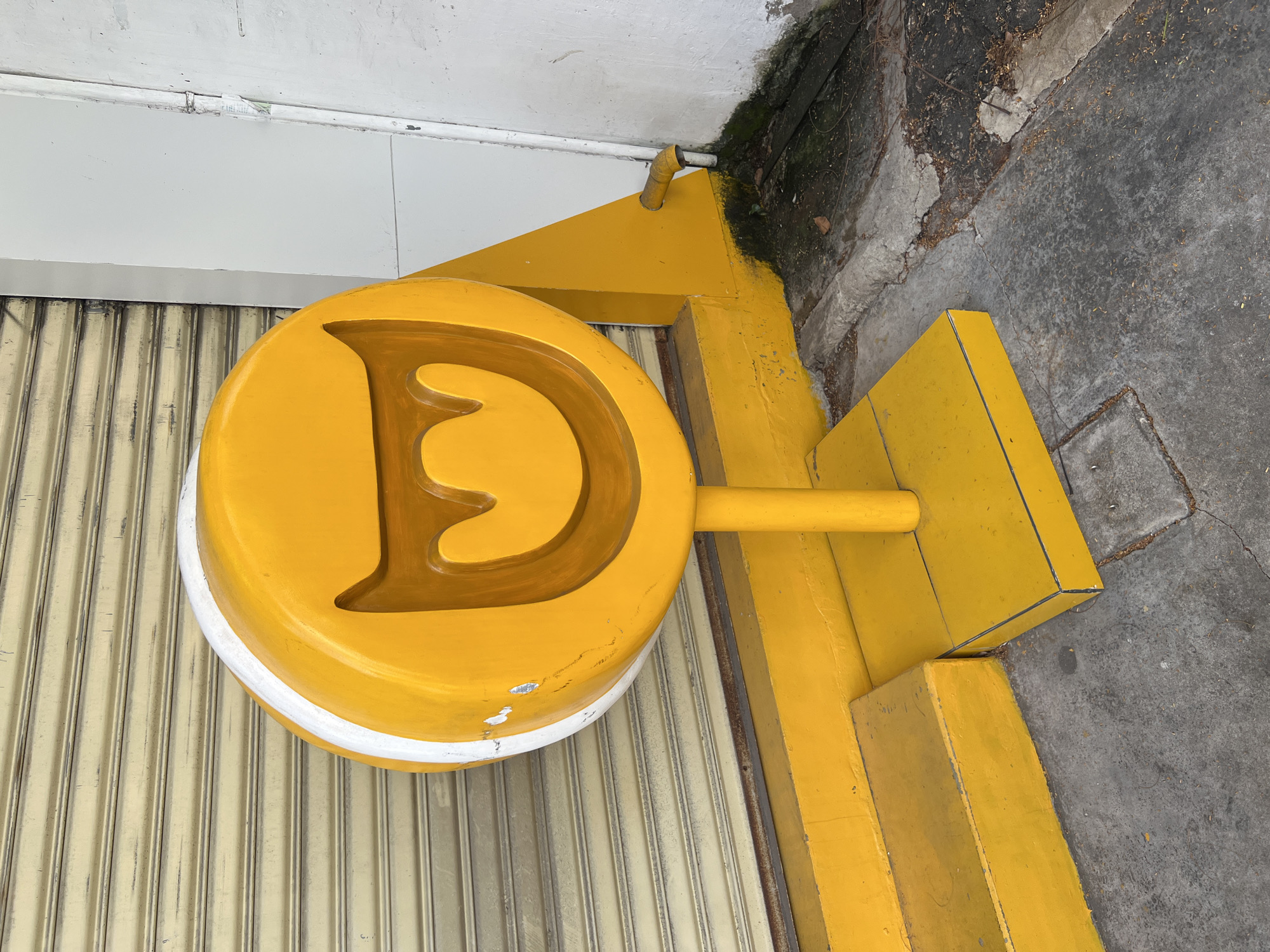 A bright yellow, circular sign with a white outline is mounted on a pole in front of a closed, beige metal storefront shutter. The sign features a brown, carved smiley face design. The sign is situated on a yellow concrete platform that extends to a yellow stepped base. To the right of the sign, a yellow angular support structure is visible, with a curved yellow drainpipe protruding from it. The ground in front of the structure is concrete, with patches of dirt, leaves, and moss. The scene appears to be outdoors, possibly in an urban setting in Nha Trang, Vietnam, given the architectural style and the presence of a drainpipe. There are no people or animals present in the image. The lighting suggests it is daytime.
