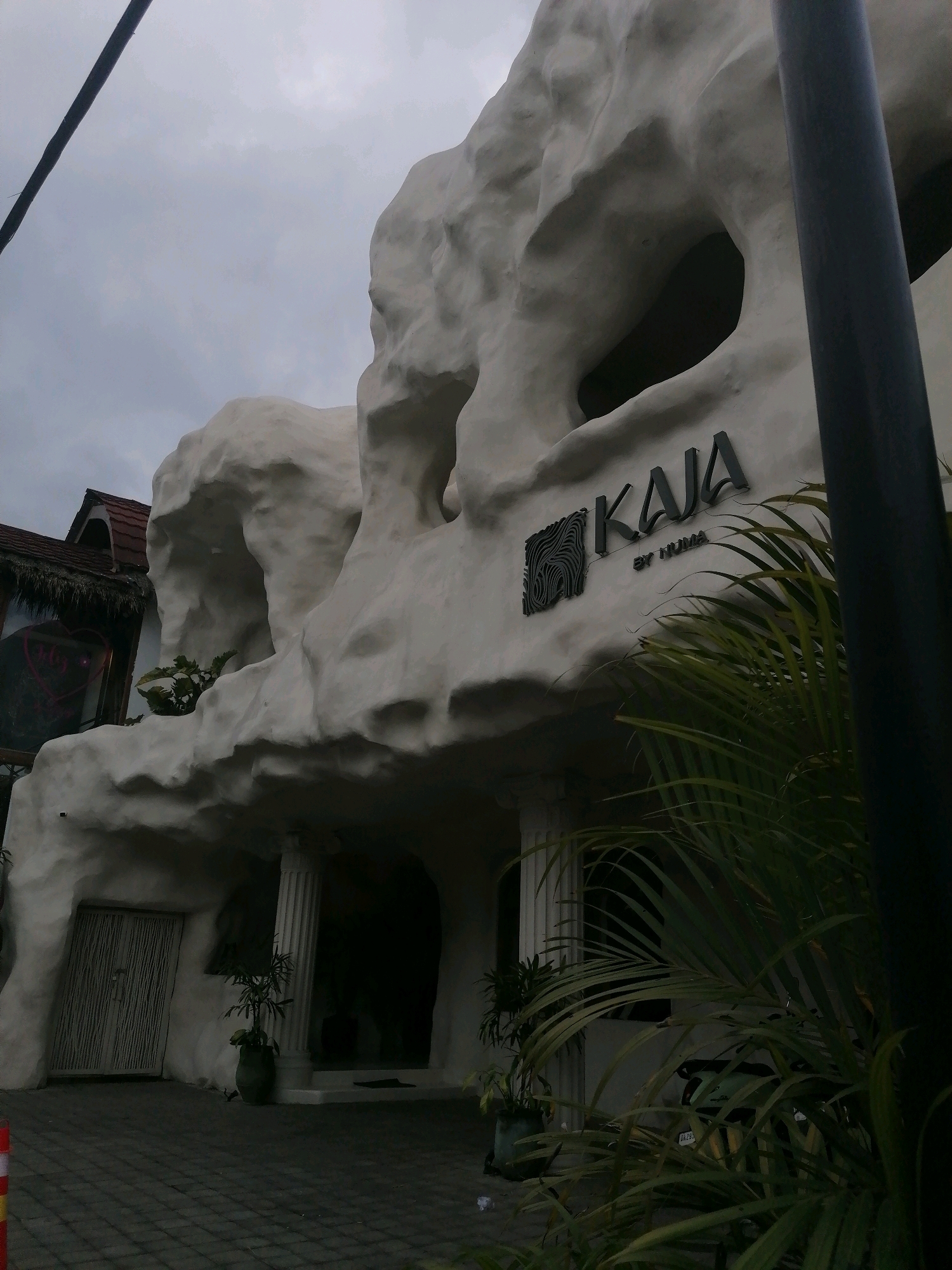 The exterior of a white, organically shaped building is visible under a cloudy sky. A dark pole is present in the foreground on the right side of the image. The building's facade features large, cave-like openings and a sign that reads "KAJA BY NUMA." A wooden door with vertical slats is on the left, and a large potted plant is situated near a pillar. In the background, a portion of another building with a thatched roof is visible. A motorcycle is partially obscured by foliage on the right side of the image. The ground in front of the building is paved with dark interlocking stones. The scene appears to be in Ubud District, Indonesia, given the architectural style and vegetation.