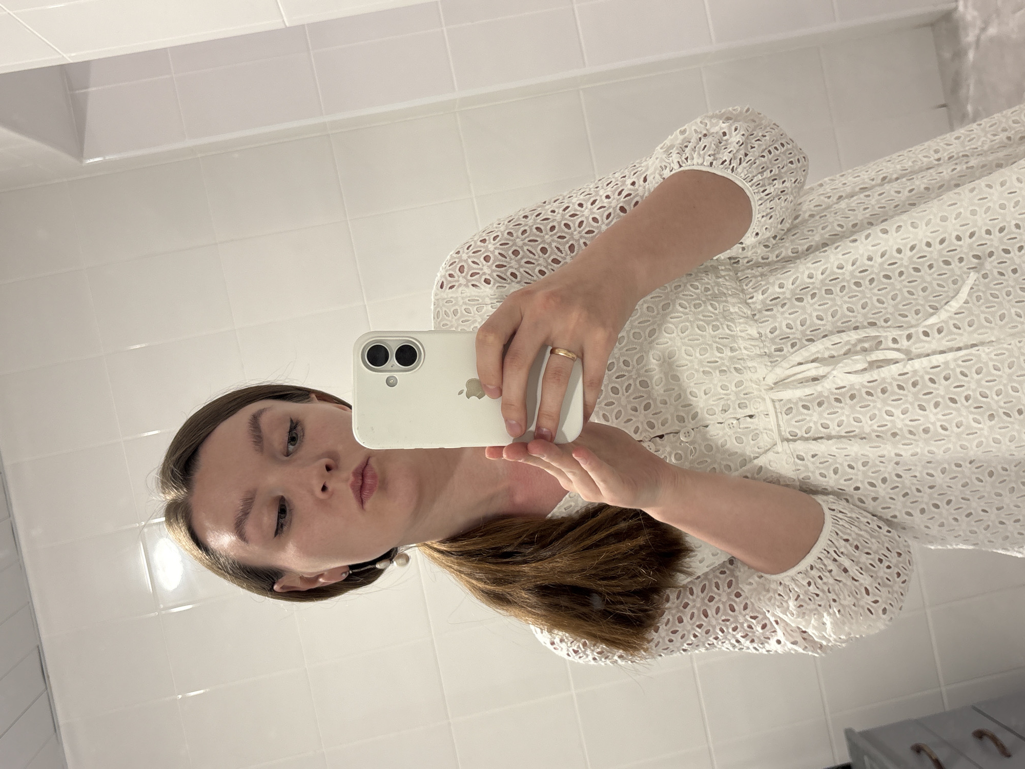 A woman is taking a selfie in a bathroom with white tiled walls. She is holding a white iPhone with two rear cameras. The woman has brown hair and is wearing a white dress with eyelet details. She is wearing a gold ring on her left hand. The location is a bathroom with white tile, but there is no clear indication that it is in Izhevsk, Russia.