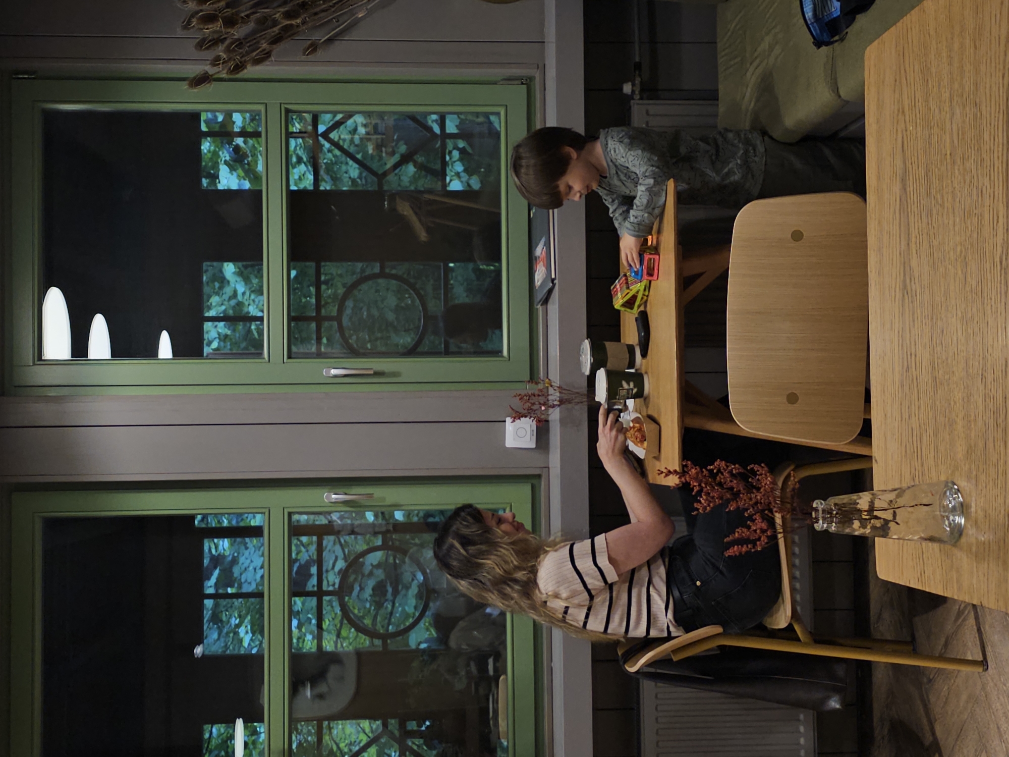 A medium shot captures a woman and a young boy sitting at separate tables inside a dimly lit establishment, possibly a cafe or restaurant, with large green-framed windows offering a view of lush foliage. The woman, seated at a table on the left, wears a striped t-shirt and dark pants. She is engaged with something on her table, possibly food, as she holds a utensil. In front of her are two disposable cups. To her right, a small bouquet of dried red flowers in a glass vase sits on a wooden table. The boy, seated at a table on the right, is dressed in a long-sleeved shirt and dark pants. He is actively playing with a colorful toy construction set on his table. Two disposable cups are also present on his table, along with a black object that could be a mobile phone. The setting appears to be in Nemchinovo, Russia, as indicated by the prompt. The overall atmosphere is calm and focused, with both individuals engrossed in their respective activities.