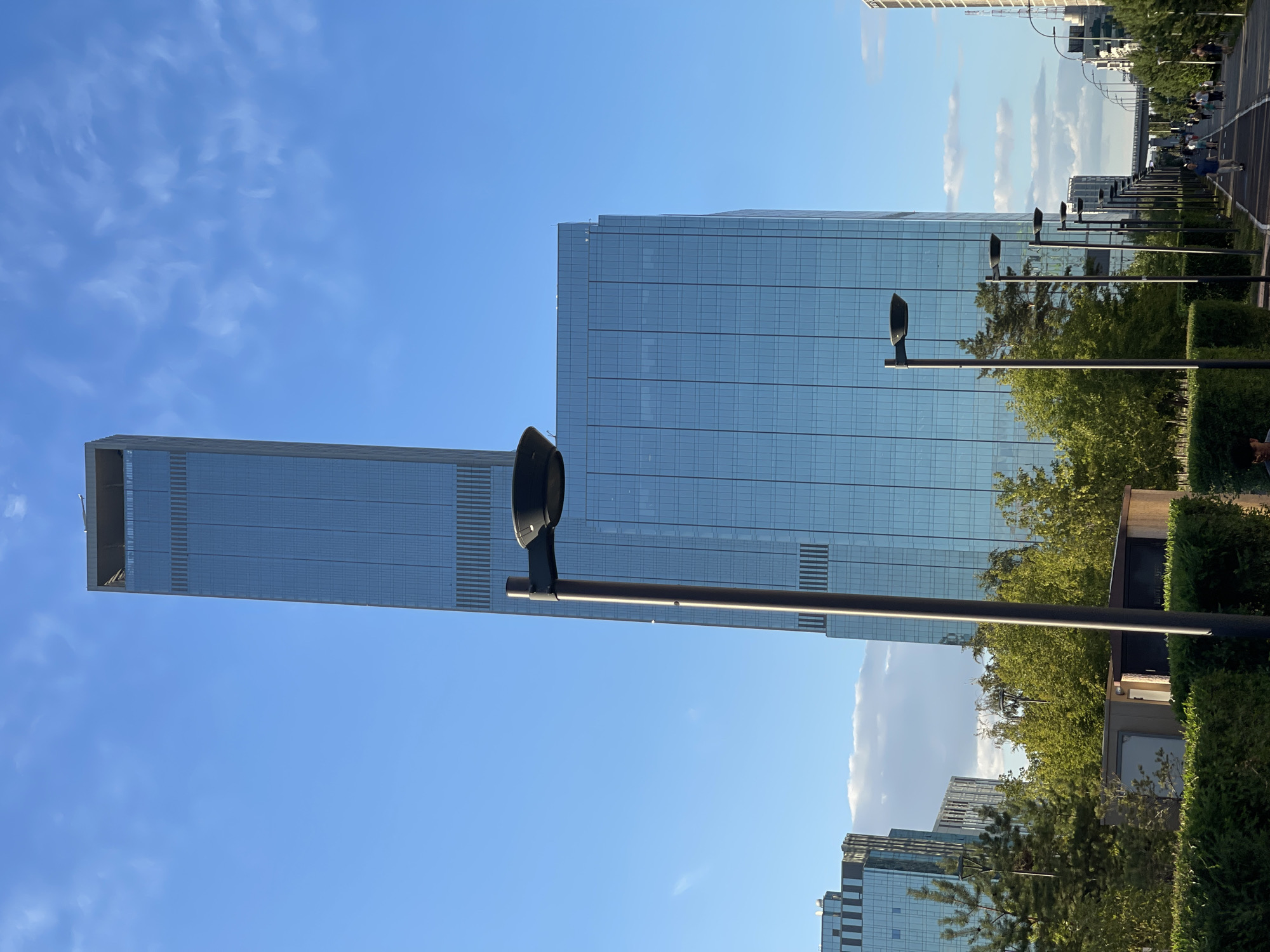 The image depicts a modern cityscape with contemporary architecture under a clear blue sky with scattered clouds. Dominating the scene are two tall, glass-clad skyscrapers, reflecting the sky. In the foreground, a paved pathway runs alongside manicured hedges and lush green trees. Several streetlights with sleek, dark lamps are strategically placed along the pathway. A few people are visible walking and strolling along the path in the distance, indicating a public outdoor space. The overall impression is one of urban development and an active public area. The scene is consistent with the modern districts of Astana, Kazakhstan, known for its futuristic architecture.