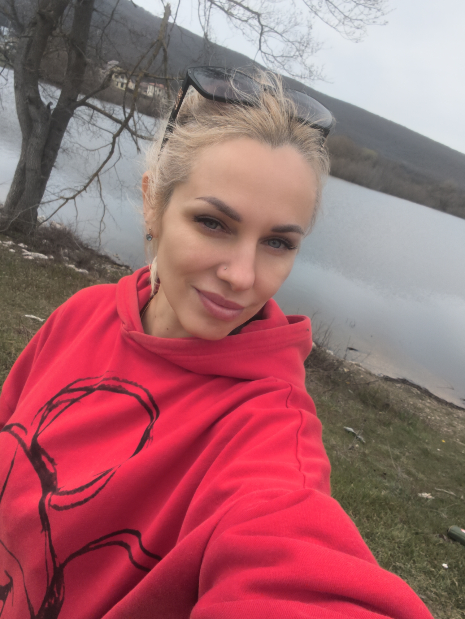 A woman is taking a selfie outdoors. She has fair skin, blonde hair pulled back, and is wearing a red hoodie. Sunglasses are perched on her head. She has blue eyes and is smiling slightly. The background consists of a lake, a tree without leaves, and a hill. The location appears to be near Орлиновский округ, Ukraine, based on the landscape and presence of the lake. The weather is overcast. The ground has patches of grass and dirt.