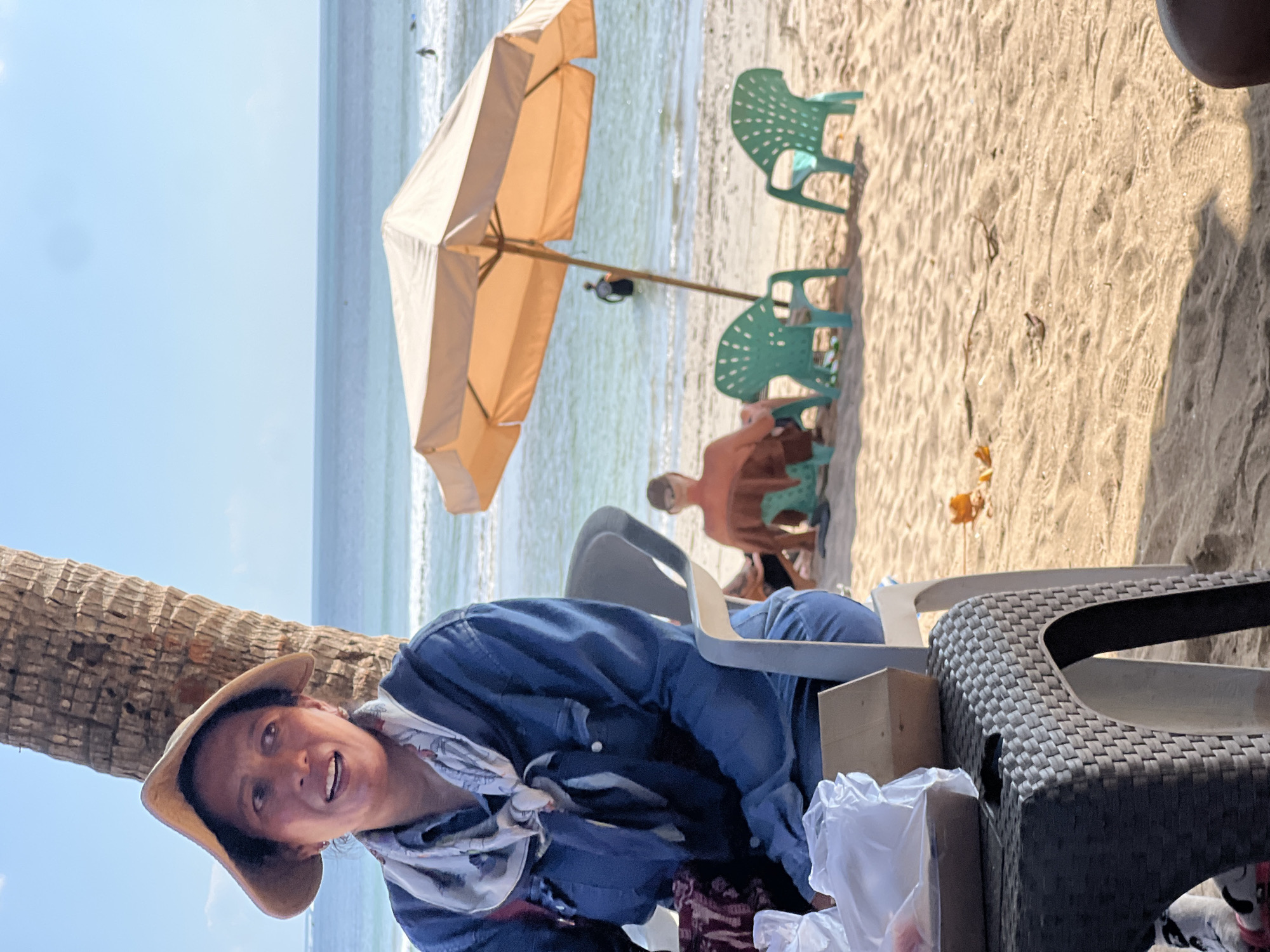 A medium shot captures a woman wearing a wide-brimmed hat and a blue denim jacket with a patterned scarf around her neck. She is seated on a chair under a palm tree on a sandy beach. Her mouth is slightly open as if she is speaking. In the background, the ocean stretches out to the horizon, with a few people visible in the water. An orange umbrella shades two green chairs on the sand. A man in an orange shirt sits facing away from the camera, and further back, another person stands in the water. A table with food items in plastic bags is in the foreground. The scene is set in Kuta, Indonesia.