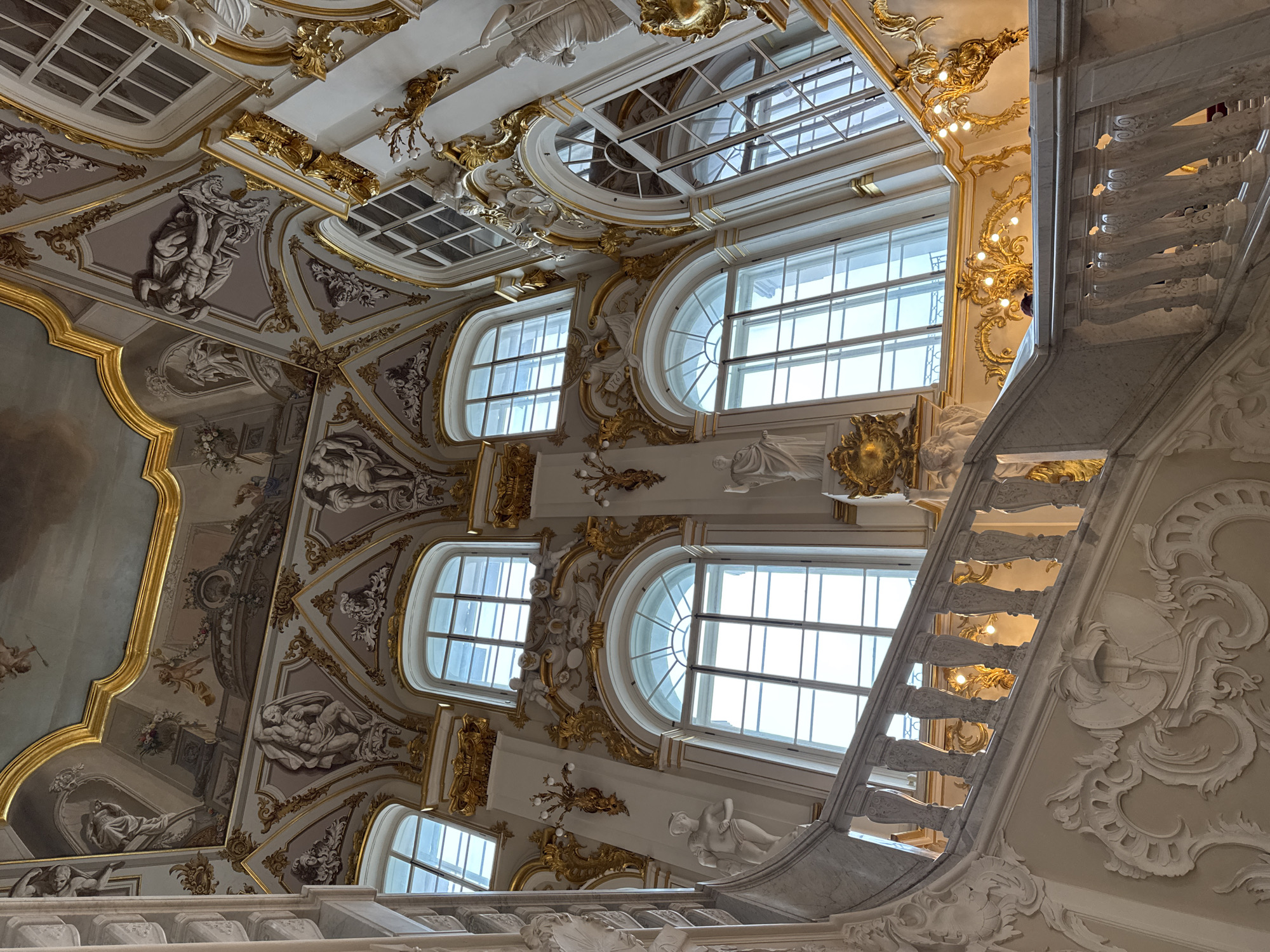 The image depicts the opulent interior of the Jordan Staircase in the Winter Palace, part of the Hermitage Museum in Saint Petersburg, Russia. The scene is dominated by a grand marble staircase with ornate white balusters and intricate white stucco moldings on the walls. Sunlight streams through tall, arched windows with multiple panes, illuminating the gilded decorative elements that adorn the walls and ceiling. These golden embellishments feature elaborate scrollwork, floral motifs, and sculptures of putti and mythological figures. The ceiling is richly decorated with frescoes and stucco reliefs, showcasing further intricate artistry. Statues of classical figures are visible on pedestals along the staircase and on the upper walls. The perspective is upward, emphasizing the grandeur and height of the space.
