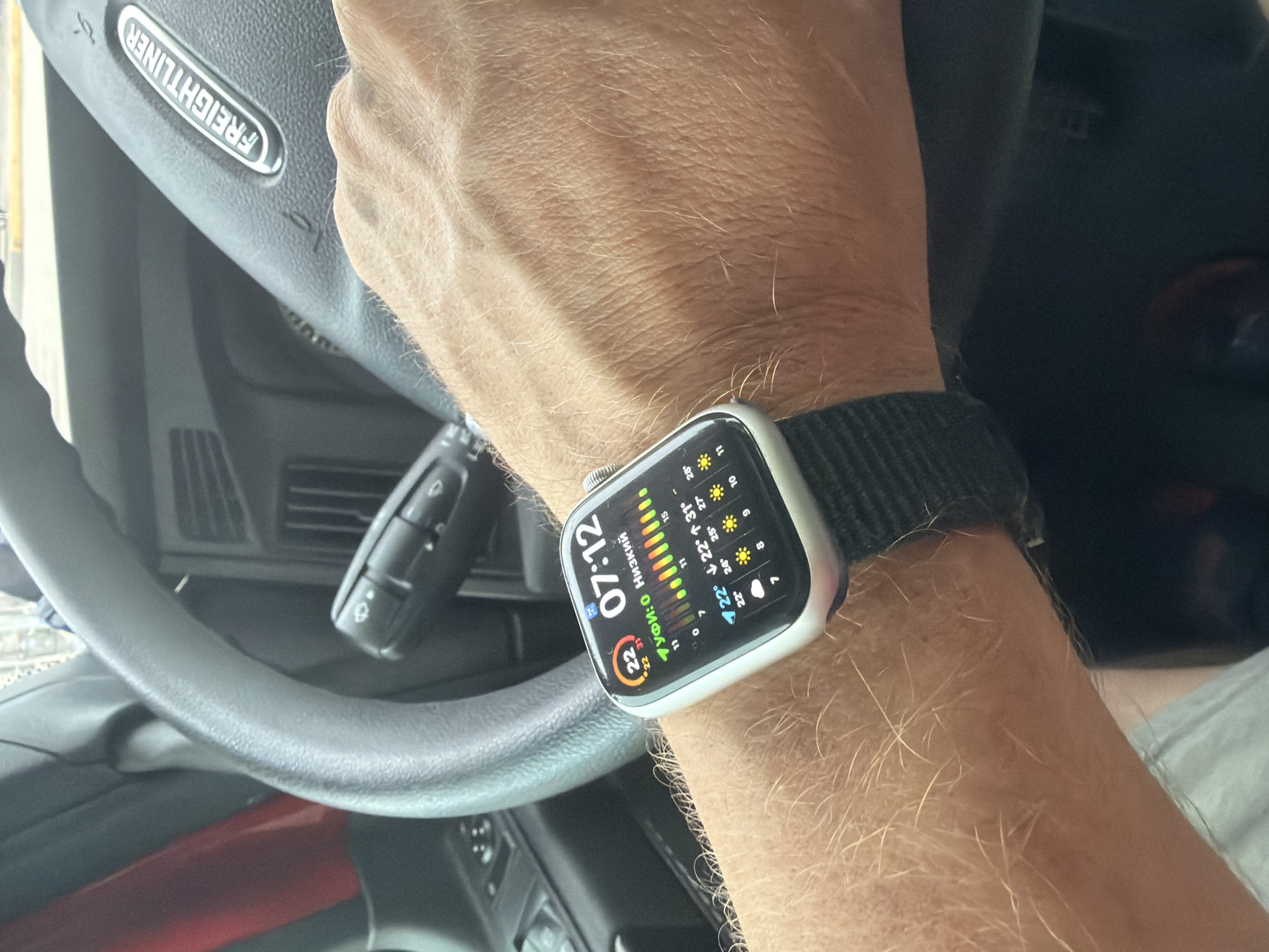 In Wildwood, United States, an individual is driving a Freightliner truck, as indicated by the logo on the steering wheel. The subject's left arm, adorned with a smartwatch displaying the time "07:12" and weather information, rests on the steering wheel. The smartwatch shows a temperature of 22 degrees Celsius and forecasts for the upcoming days, with icons depicting sunshine and varying degrees of cloudiness. The environment appears to be an interior of a vehicle, likely a truck cab, with the steering wheel and dashboard visible. The overall lighting suggests it is daytime.