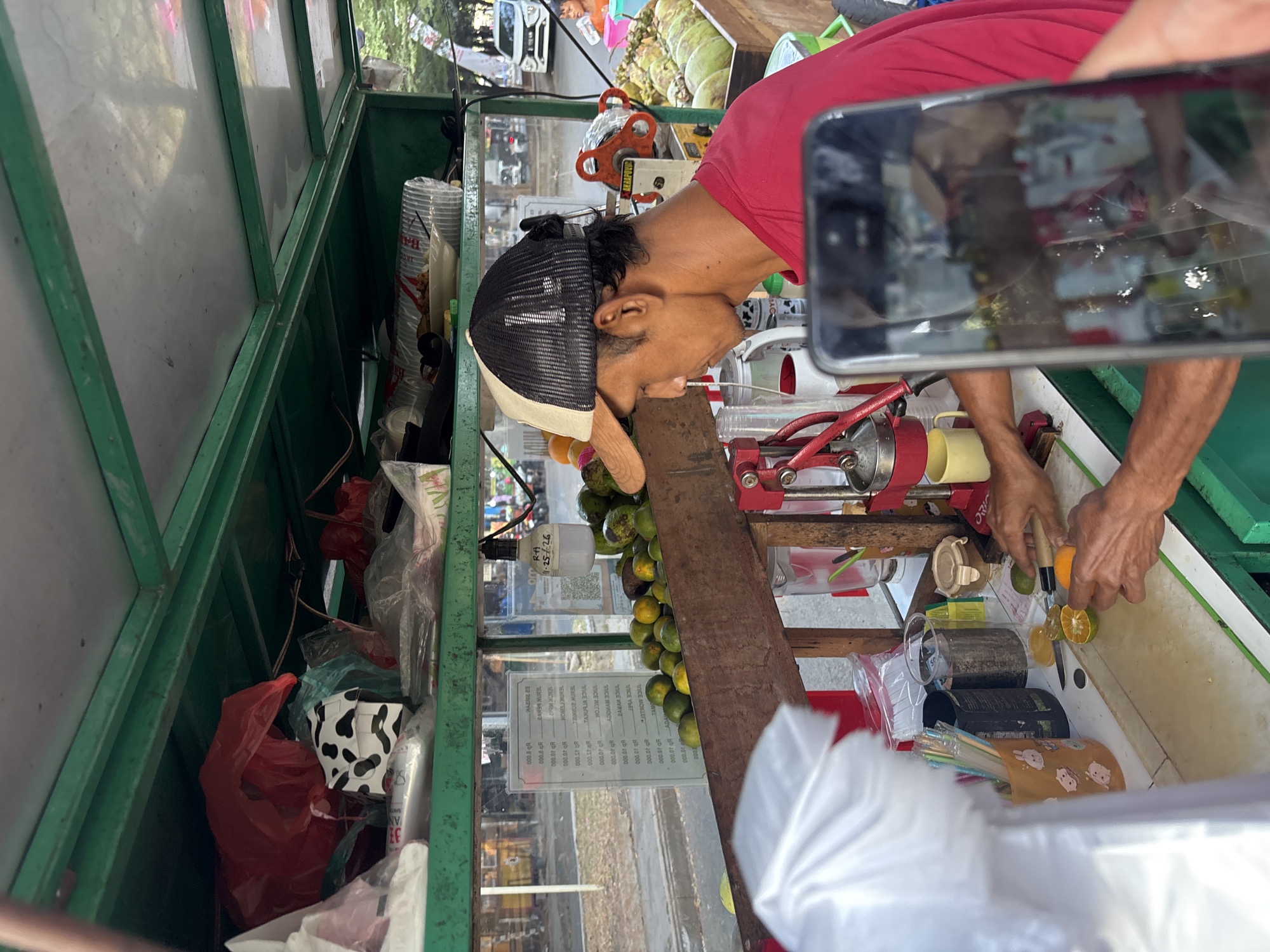 The image depicts a street vendor preparing fresh juice in Cirebon, Indonesia. A man wearing a red shirt and a two-toned cap is seen behind a cart filled with fruits, primarily oranges and coconuts. He is in the process of cutting oranges with a knife on a white cutting board, with a manual citrus juicer beside him. The cart is stocked with various items, including plastic cups, straws, and what appear to be plastic bags for packaging. In the background, a menu board is visible with text in Indonesian. The scene appears to be outdoors, on a street, with traffic and pedestrians visible in the distance. The overall impression is that of a bustling, everyday scene in a Southeast Asian city.