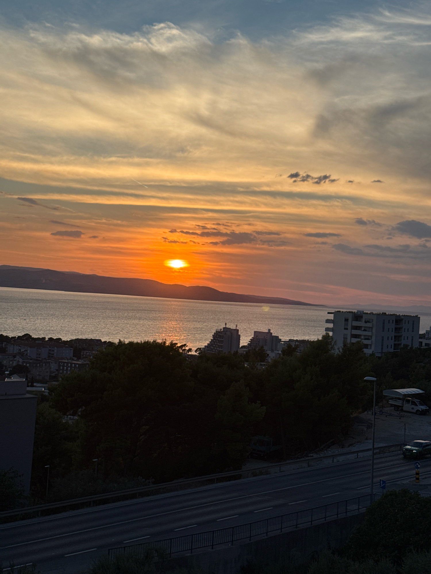 This image captures a sunset over the Adriatic Sea in Makarska, Croatia. The sky is a vibrant blend of orange, yellow, and blue, with wispy clouds streaking across the horizon. The sun, a bright orb, is setting behind a silhouetted mountain range in the distance. In the foreground, a body of water, likely the sea, reflects the warm hues of the sunset. Along the coastline, there are buildings and trees, indicating a populated area. A highway with cars is visible in the lower portion of the image, suggesting urban activity. The overall scene is serene and picturesque, showcasing the natural beauty of the Croatian coast at dusk.