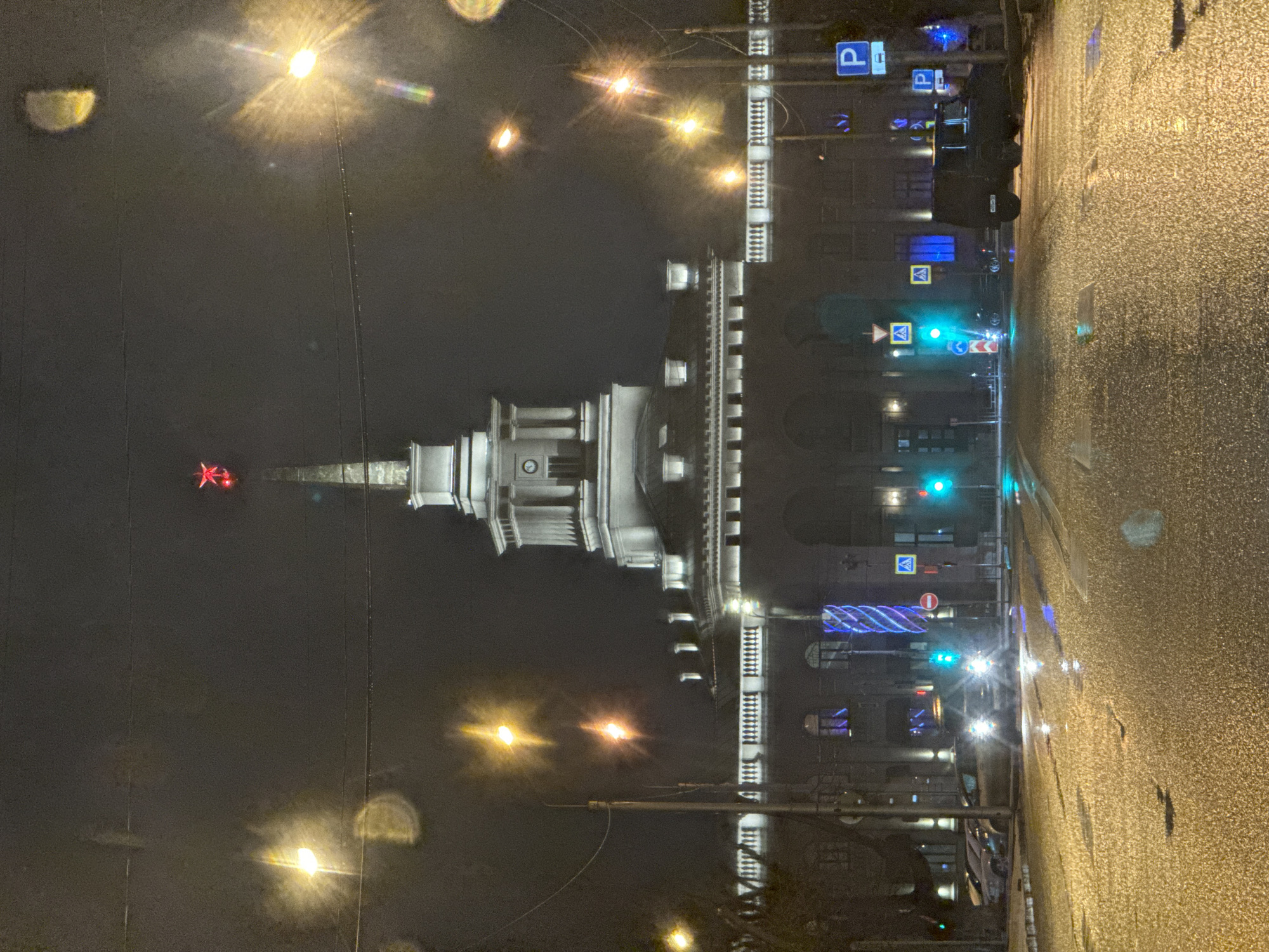 At night in Krasnoyarsk, Russia, the photograph shows a wet, paved road alongside a building with a tower topped by a red star. Streetlights illuminate the scene, creating glare and reflections on the wet surface. Traffic lights can be seen along the street. The building is white and lit with architectural lighting. There are no people visible in the photograph.
