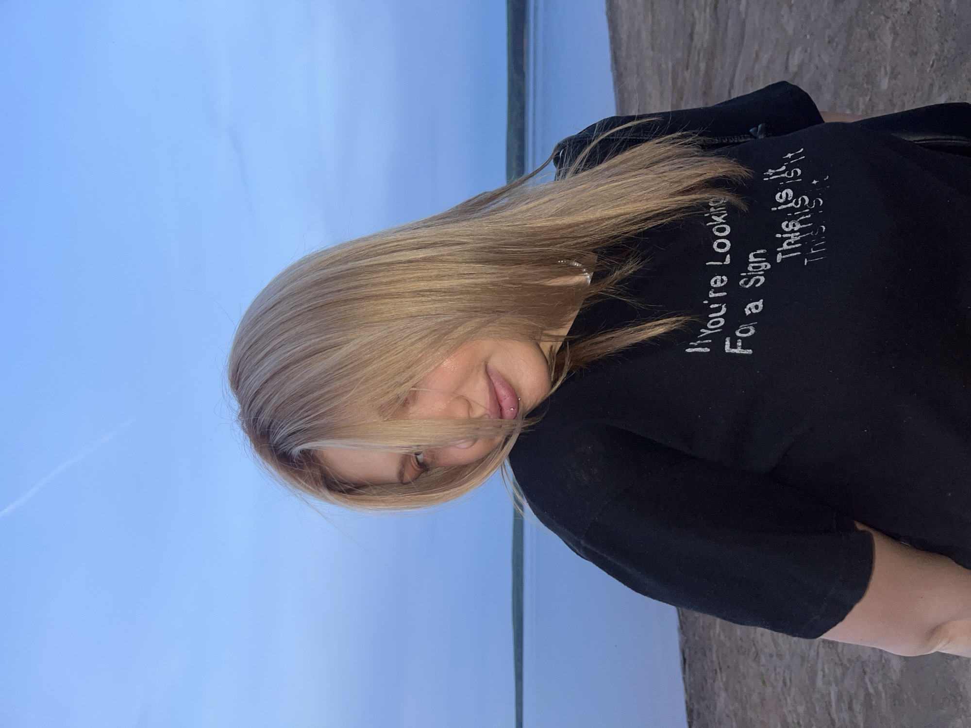 A young woman with blonde hair stands outside near a body of water under a pale blue sky, possibly during the early evening. She is wearing a black long-sleeved shirt with white text on the back. The text says, "If You're Looking For a Sign, This is it This is it". The background appears to be a river and its shore in Chaikovsky, Russia. The woman is angled slightly towards the camera, her face partially obscured by her hair.