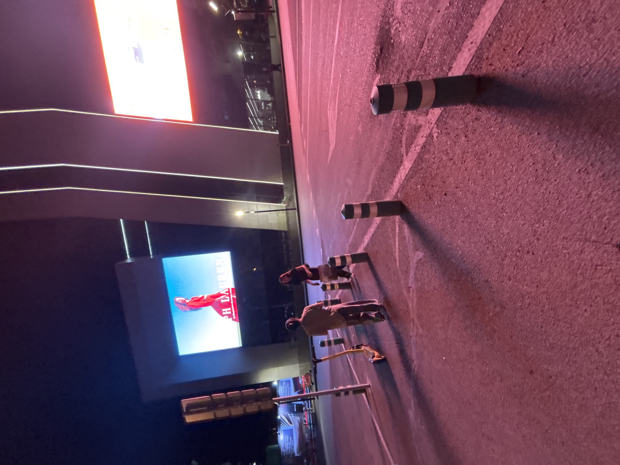 At night in Tbilisi, Georgia, two individuals stand on a road illuminated by pinkish-purple light. To the left, a yellow electric scooter is parked near a traffic light pole. A large LED screen is mounted on a building in the background, displaying an advertisement with a woman in a red dress and the text "PH LAUREN". To the right, another large screen glows with a red and white advertisement. The road surface is asphalt, marked with white lines. Several bollards with black and white stripes are placed along the road. The scene appears to be an urban environment, possibly a public square or a commercial area.