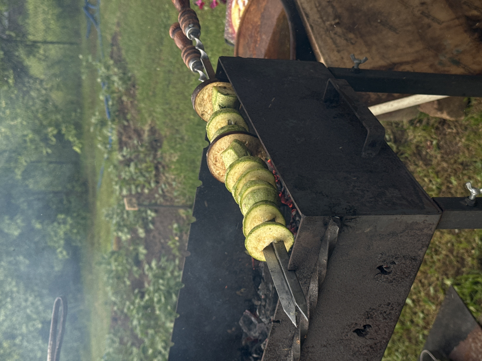 In an outdoor setting, likely a backyard or garden, possibly in the vicinity of Поназырево, Russia, a metal barbecue grill is filled with glowing coals. Skewers laden with alternating slices of zucchini and eggplant are positioned horizontally over the heat, indicating food preparation. Smoke rises from the grill, suggesting active cooking. The background reveals green foliage and trees, consistent with a rural or suburban environment. The overall scene captures a moment of outdoor cooking.
