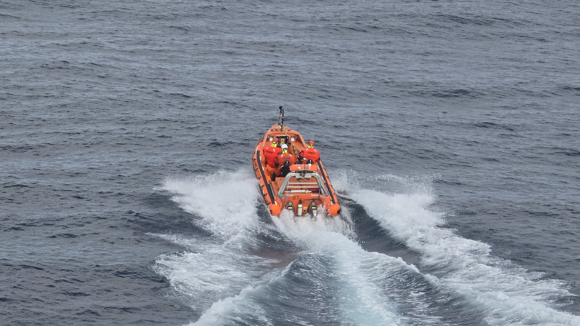 An orange inflatable boat, occupied by four individuals in red suits, is moving rapidly through the ocean. The boat leaves a white wake behind it. The location is not specified.
