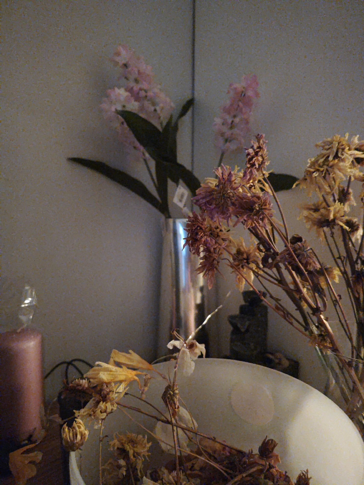 The image depicts a still life arrangement of flowers and a candle in an indoor setting. In the foreground, a tall, pink candle is visible on the left, next to a white, egg-shaped object. Behind these items, dried flowers in shades of yellow and purple are arranged in a vase. A portion of a metallic vase holding what appear to be pink and green artificial flowers is visible in the background, to the left of the dried flowers. The background wall has a textured, speckled appearance and is a neutral, light color. The lighting is dim, suggesting an indoor environment with artificial light. There are no people or animals present in the image. The scene is static and appears to be a decorative display.