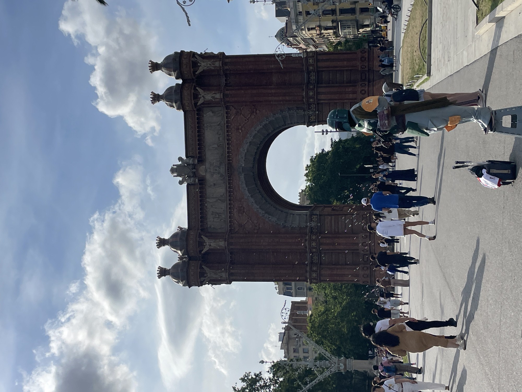 The image depicts a bustling outdoor scene in front of the Arc de Triomf in Barcelona, Spain. The prominent central feature is the reddish-brown brick triumphal arch, adorned with three crown-like finials and various sculptural reliefs, under a partly cloudy blue sky. Numerous small bubbles are visible floating in the air, particularly near the right side of the arch.
In the foreground, slightly right of center, a street performer dressed as the Star Wars character Boba Fett stands stationary on a grey plastic stool. The costume is green and grey with orange elements, and the performer holds a prop rifle. A small black wheeled suitcase with a red and white item attached rests on the ground beside the stool. A person's legs and lower torso are partially visible behind the performer.
A wide, paved pedestrian area extends from the foreground towards the arch. Many individuals are walking across this area in various directions. On the left, a woman in a light brown dress and white shoes walks away from the viewer, alongside a person in a white t-shirt and black pants. Further left, a woman in a white top and white trousers walks towards the viewer. On the right, a man in a blue t-shirt and white cap walks away from the viewer, as does a woman in a long white shirt and shorts. Shadows of pedestrians are cast onto the paved ground.
Trees and green foliage line both sides of the thoroughfare, with more dense greenery visible behind the arch. Ornate lampposts are present on the left and right sides of the frame. Buildings with traditional architecture, including balconies and domes, are visible in the background on the right side of the image.
