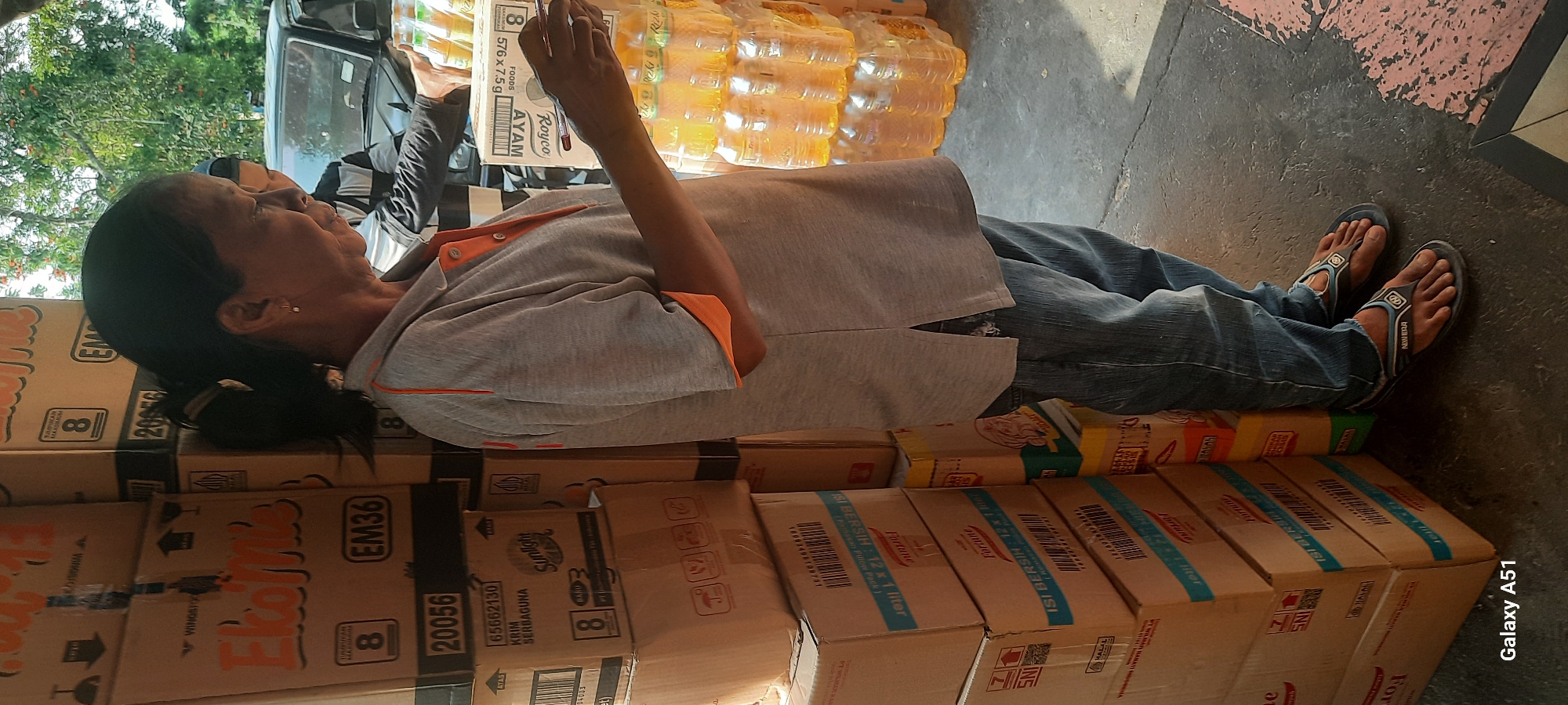 In the city of Beru, Indonesia, a woman wearing a grey polo shirt with orange trim and blue jeans stands next to stacks of cardboard boxes. She is holding a package of bottled beverages, likely palm oil, and appears to be arranging them or checking them. The boxes are labeled with various product names and numbers, suggesting a commercial setting, possibly a market or warehouse. In the background, a person in a black and white striped shirt is visible behind the woman, also interacting with the beverages. A dark-colored vehicle is partially visible in the background, with a portion of its front end showing. The ground is paved, and sunlight casts shadows, indicating an outdoor or semi-outdoor environment. The overall scene depicts a moment of commerce or inventory management.