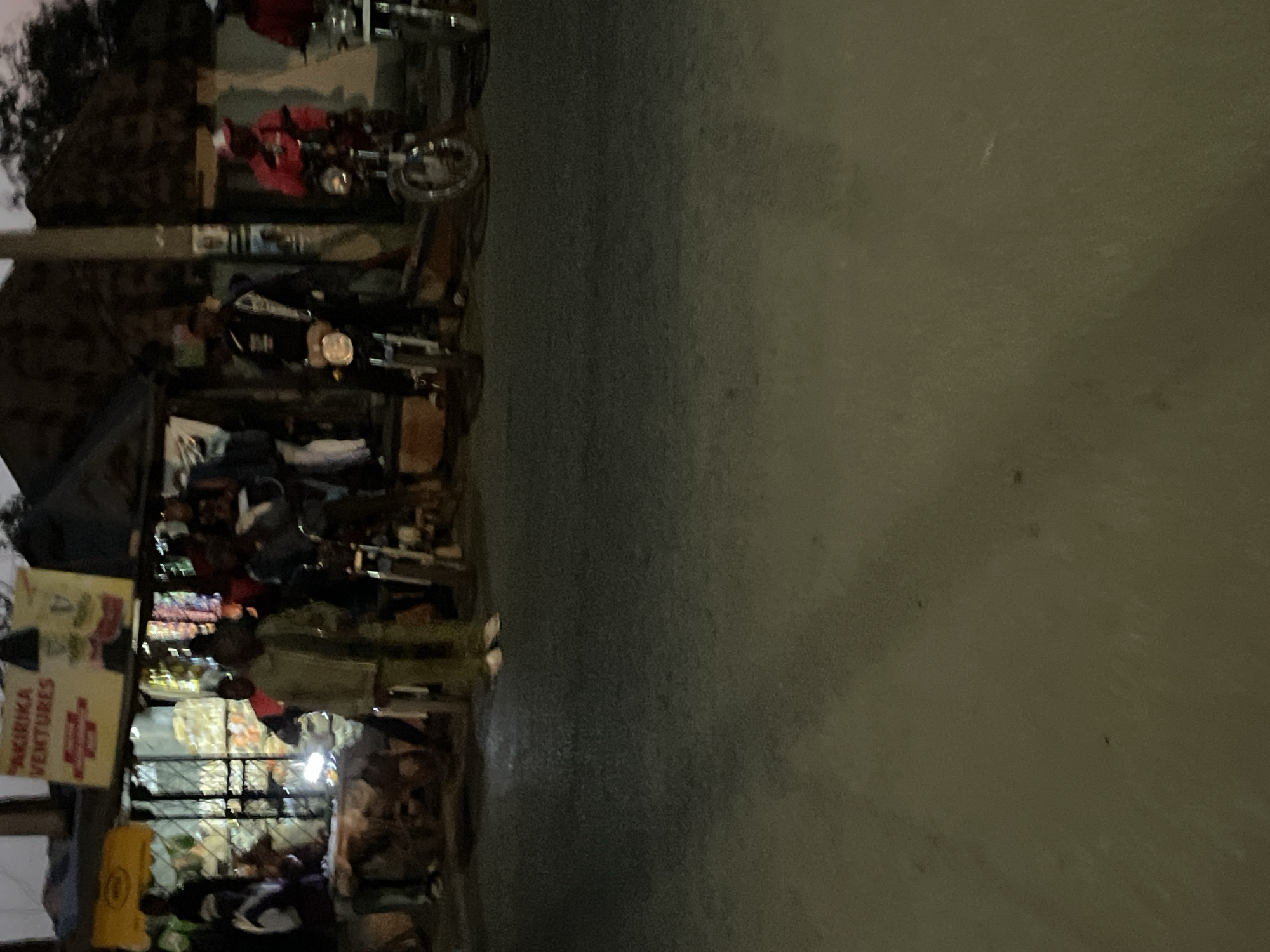 The image shows a roadside scene in Bukuru, Nigeria, at dusk. A small shop or kiosk identified as "Akirika Ventures" is visible. People are gathered around, likely customers. A motorcycle is parked nearby. The road is dark, suggesting low lighting. Other businesses and people are visible in the background but are out of focus. The overall atmosphere is typical of a busy street in a developing urban area.