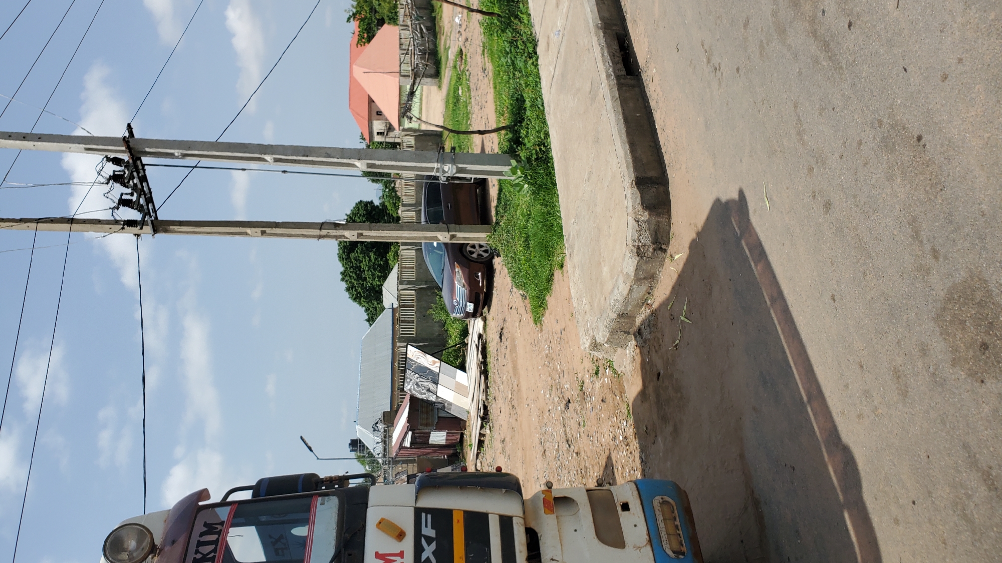 The image shows a street scene in Jalingo, Nigeria. On the left, the front of a white truck with "KIM" visible on its side is parked. In the middle ground, a brown SUV is parked near a display of tiles. The background features several buildings with corrugated metal and tiled roofs, some with concrete walls. Overhead, power lines and a utility pole are prominent. The ground is a mix of dirt and pavement, with a concrete curb in the foreground. The sky is partly cloudy.