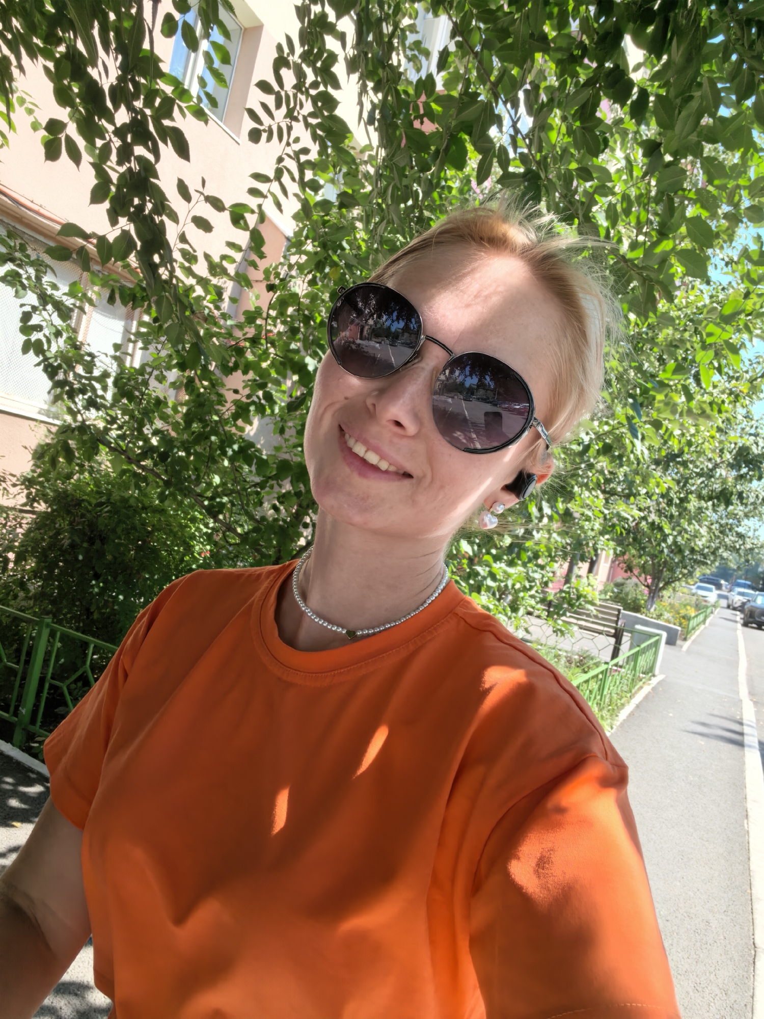 In an outdoor, street-level, medium shot, a blonde woman with fair skin is taking a selfie. She is wearing an orange t-shirt, a silver necklace, dark round sunglasses, and black earbuds with pearl-shaped earrings. Her head is tilted slightly to her left, and she is smiling. In the background, there is a pale pink building with windows, green foliage from trees and bushes, a green metal fence, and a paved sidewalk with cars parked on the street. The lighting suggests it is daytime with sunlight filtering through the leaves, creating dappled shadows on the woman and her shirt. The location is indicated to be Sterlitamak, Russia.
