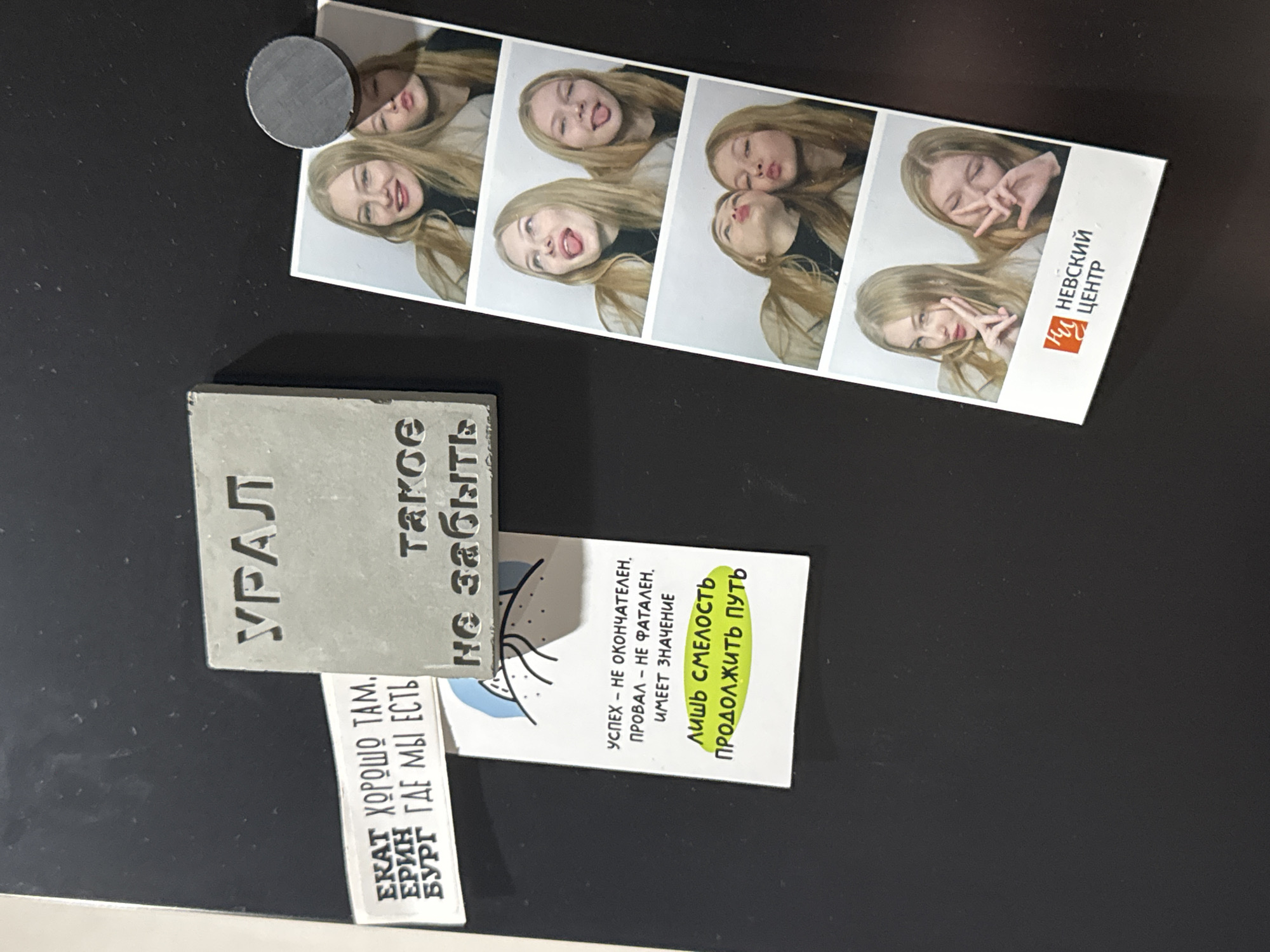 The image depicts a collection of items affixed to a dark, possibly black, surface. The items include: A photo strip featuring a young woman with blonde hair making various silly faces in four successive frames. A grey, square tile with the word "УРАЛ" (URAL) embossed in Cyrillic letters. A small white strip with the phrase "ЕКАТ ХОРОШО ТАМ. БУРГ ГДЕ МЫ ЕСТЬ" (EKAT IS GOOD THERE. BURG WHERE WE ARE) printed on it. A card with text that includes "УСПЕХ - НЕ ОКОНЧАТЕЛЕН, ПРОВАЛ – НЕ ФАТАЛЕН, ИМЕЕТ ЗНАЧЕНИЕ" (SUCCESS - NOT FINAL, FAILURE - NOT FATAL, IT HAS MEANING) and "ЛИШЬ СМЕЛОСТЬ ПРОДОЛЖИТЬ ПУТЬ" (ONLY COURAGE TO CONTINUE THE PATH). A magnetic disc is attached to the photo strip. The photo strip also bears the inscription "НЕВСКИЙ ЦЕНТР" (NEVSKY CENTER). The overall impression is that these are keepsakes or souvenirs. It is impossible to pinpoint the city of Perm in Russia without additional information from this image.