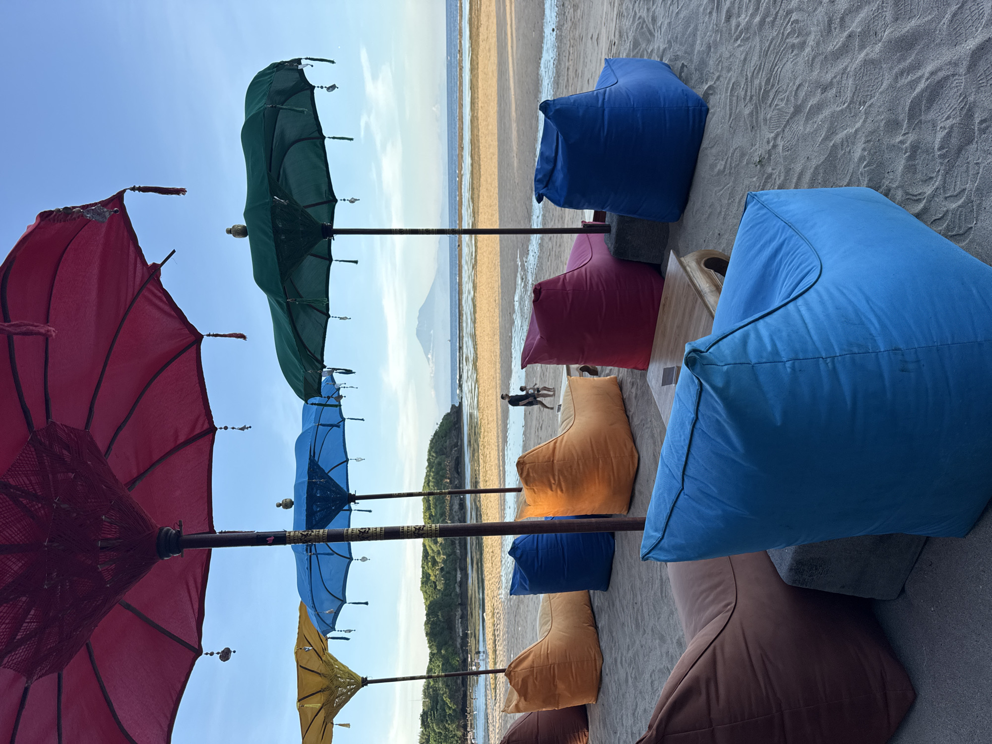 The image depicts a beach scene in Kuta Selatan, Indonesia. The foreground features a row of colorful beanbag chairs and ornate parasols in red, green, blue, and yellow shades, positioned on sandy ground. The parasols are arranged in a linear fashion, extending towards the backdrop.
The midground shows a beach area with a person and a child walking along the shoreline.
In the background, a body of water meets a sandy beach. The sky is blue. The overall lighting suggests a sunny day.