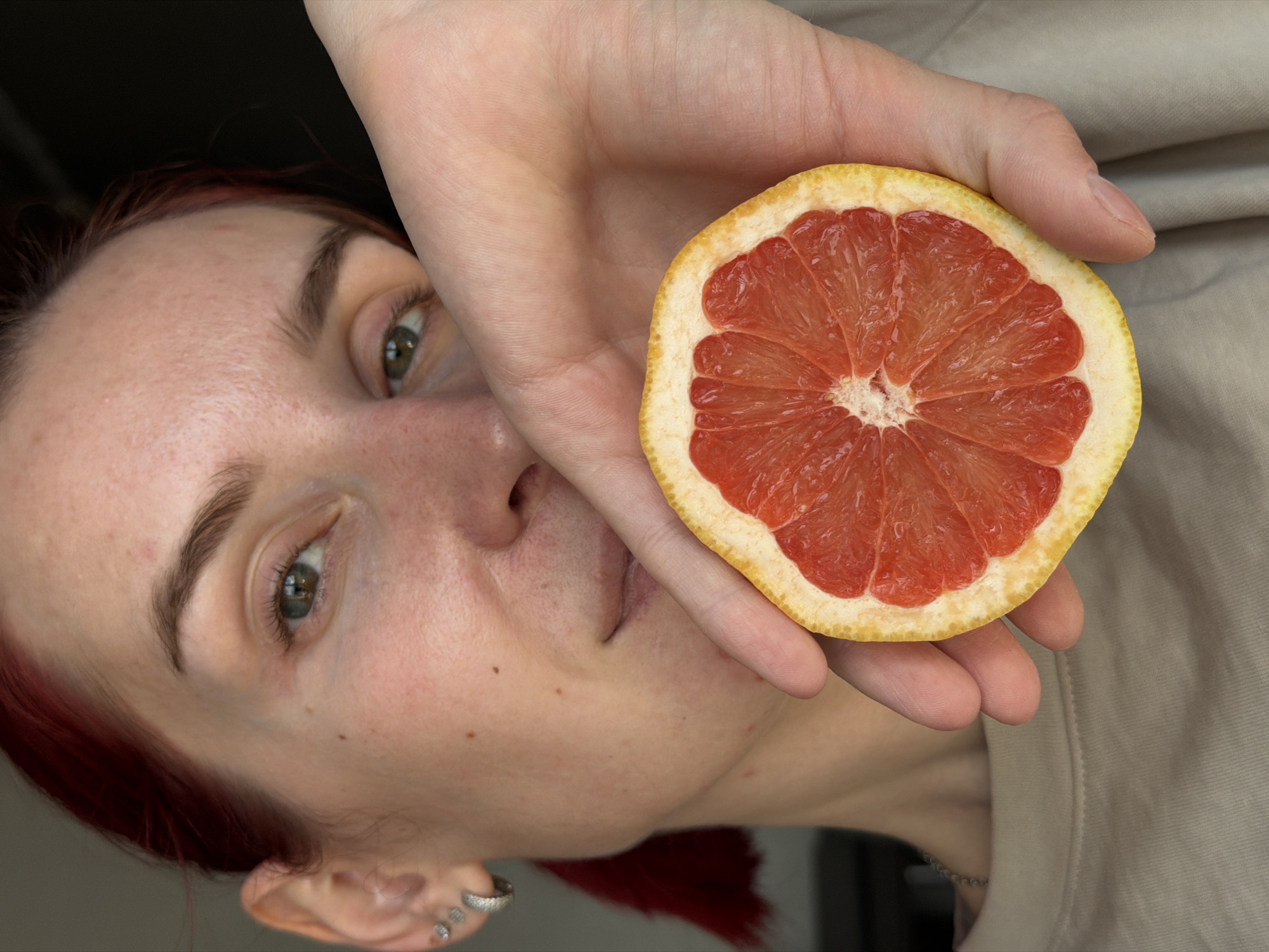 A close-up, rotated portrait shot captures a person holding half of a red grapefruit in front of their face. The subject has light skin, reddish hair visible on the left and bottom of the frame, and green-hazel eyes that look directly at the viewer. Their left eye is prominent, while the right side of their face is partially obscured by their hand holding the fruit. Distinct details on the face include visible pores and a small mole on the cheek. The left ear is adorned with multiple small rings, including helix and lobe piercings.
The vibrant red-pink segmented flesh and white pith of the grapefruit are sharply in focus, contrasting with the yellowish rind. The fruit is held in the palm of the person's hand, with fingers visible gripping the top edge. The background on the left side is dark and out of focus, while a soft, beige-colored fabric, possibly clothing, is visible on the right.
The image suggests an indoor setting in Chaikovsky, Russia, with the subject engaging the camera with a neutral expression. No specific time of day or weather conditions are discernible, and no text is visible.