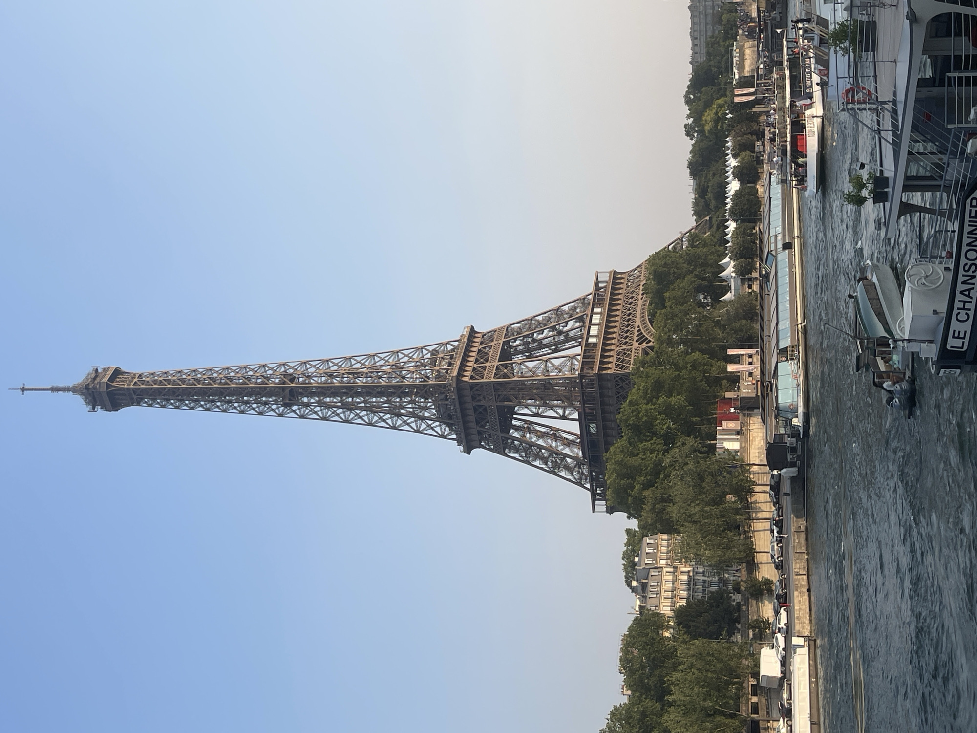 The image shows the Eiffel Tower in Paris, France, viewed from across the Seine River. The Eiffel Tower, a wrought-iron lattice tower, dominates the upper portion of the frame, with its intricate structure clearly visible against a clear blue sky. In the foreground, the Seine River flows, with several boats of various sizes on its surface. One boat, labeled "LE CHANSONNIER," is prominent in the lower right corner. To the left of the Eiffel Tower, along the riverbank, are trees, buildings, and parked cars. A large tour boat with passengers is visible in the middle ground, also on the river. The overall scene depicts a sunny day in Paris, with the iconic landmark as the central focus.