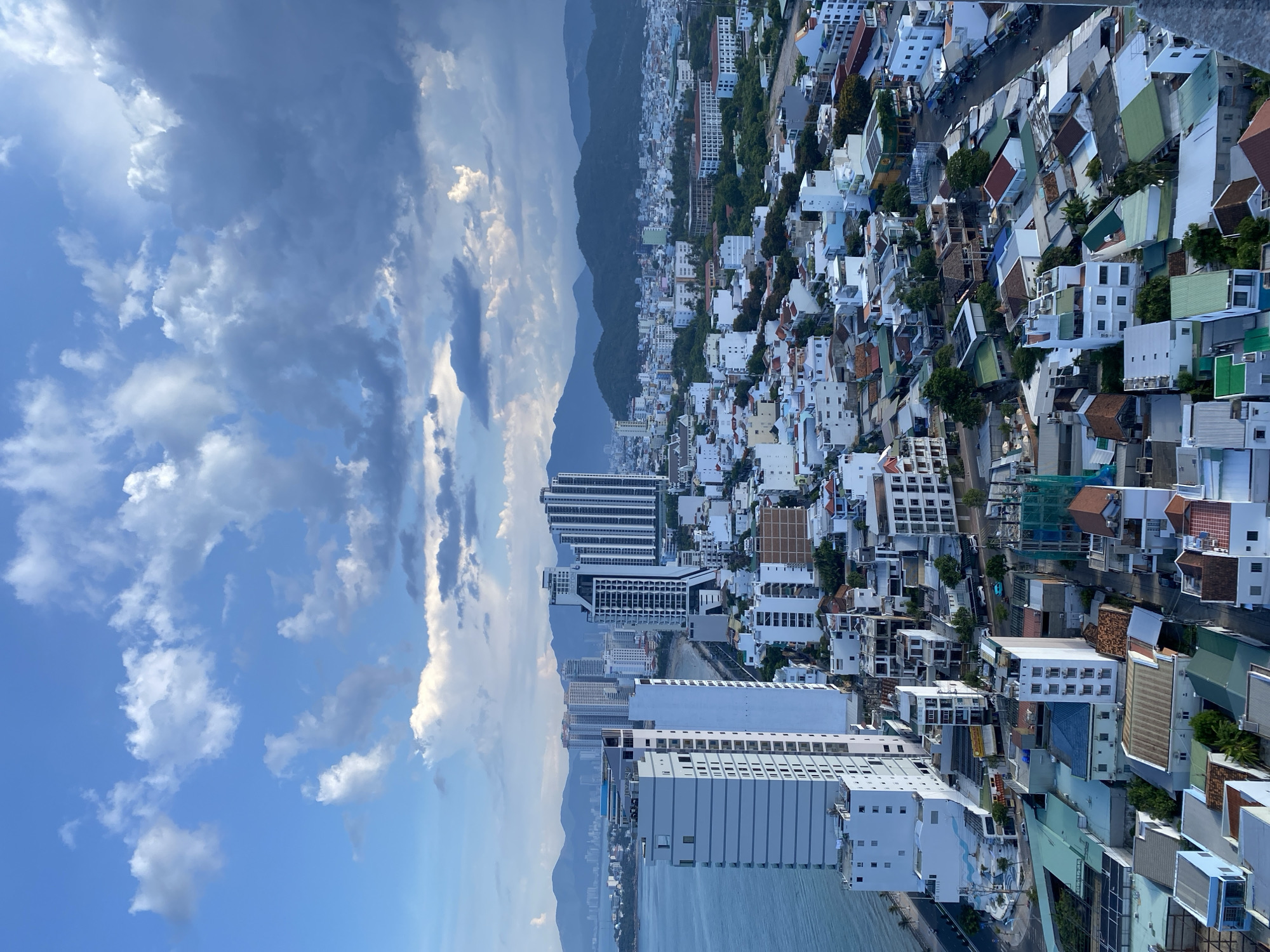 An aerial perspective captures the coastal city of Nha Trang, Vietnam, under a dramatic sky. The foreground is dominated by a dense urban landscape featuring a mix of low-rise residential buildings with diverse roofing materials, including terracotta, green, and blue, interspersed with taller, modern white and grey apartment complexes and hotels. One particularly prominent, slender white building stands out on the left side. Beyond the immediate urban sprawl, a body of water, likely the South China Sea, is visible to the left, bordered by a road and a thin strip of beach. In the background, a range of lush, green mountains forms a natural backdrop to the city, with faint signs of further development on their lower slopes. The sky is a striking feature, with patches of bright blue contrasted by dark, heavy clouds that suggest an approaching storm or recent rainfall. The scene is devoid of visible people or vehicles, presenting a static, architectural view of the city.