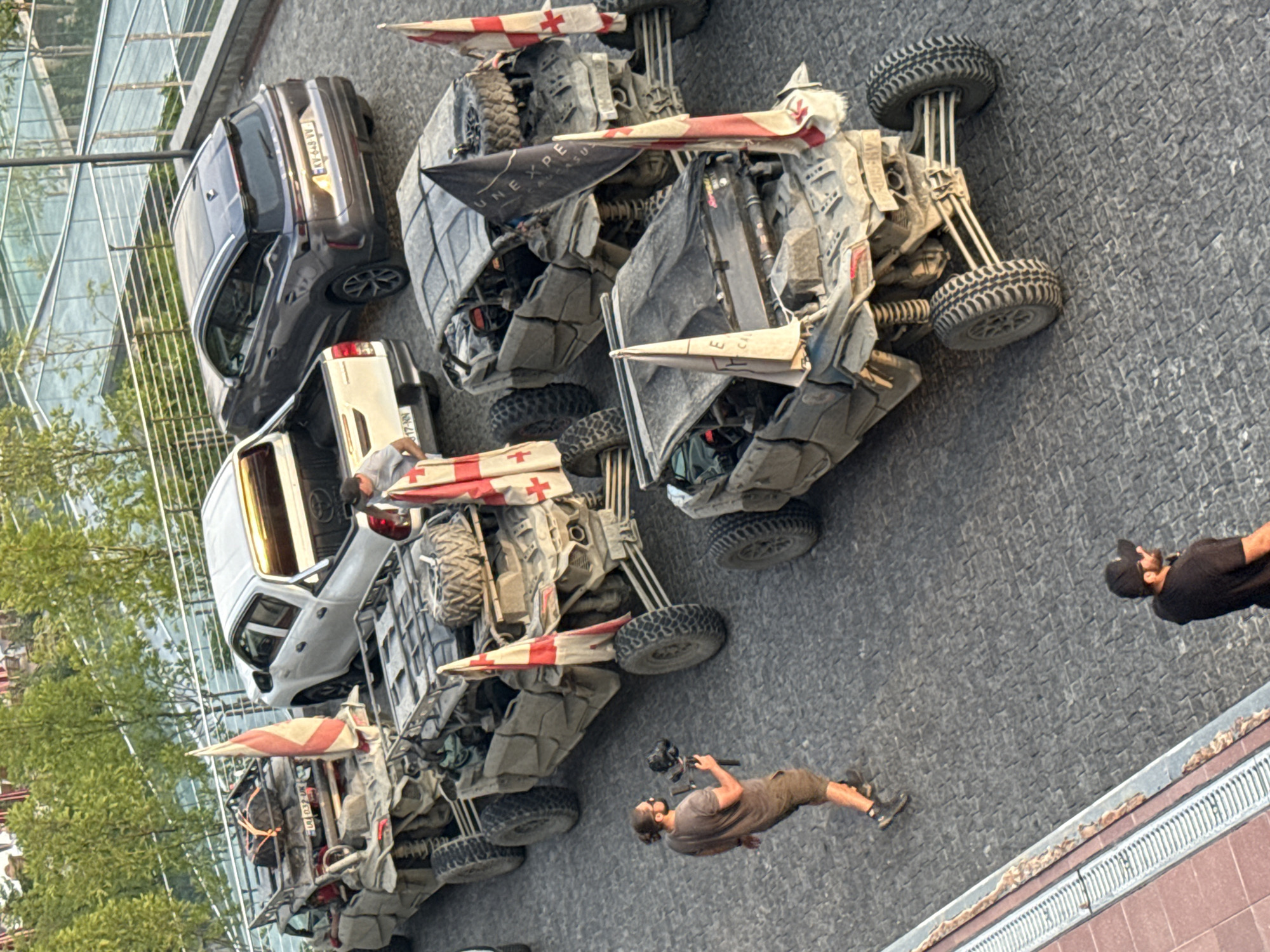 The image captures an outdoor scene in Sighnaghi, Georgia, featuring a lineup of rugged, sand-colored off-road vehicles, likely ATVs or similar recreational vehicles, parked on a cobblestone surface. Several of these vehicles are adorned with Georgian flags, indicating a local or national affiliation. In the foreground, a man with dark hair tied back in a bun, wearing a grey t-shirt and shorts, is seen walking and holding a professional-looking camera on a gimbal, suggesting he is documenting the scene or an event. Further back, another man wearing a black t-shirt and a baseball cap is standing, looking away from the camera. A white pickup truck and a dark-colored SUV are also present in the background, positioned behind the off-road vehicles. The environment appears to be a paved area with some greenery and modern architectural elements visible in the upper portion of the frame, hinting at a developed urban or tourist area. The vehicles are covered in a fine layer of dust or sand, implying recent use in a dusty environment.