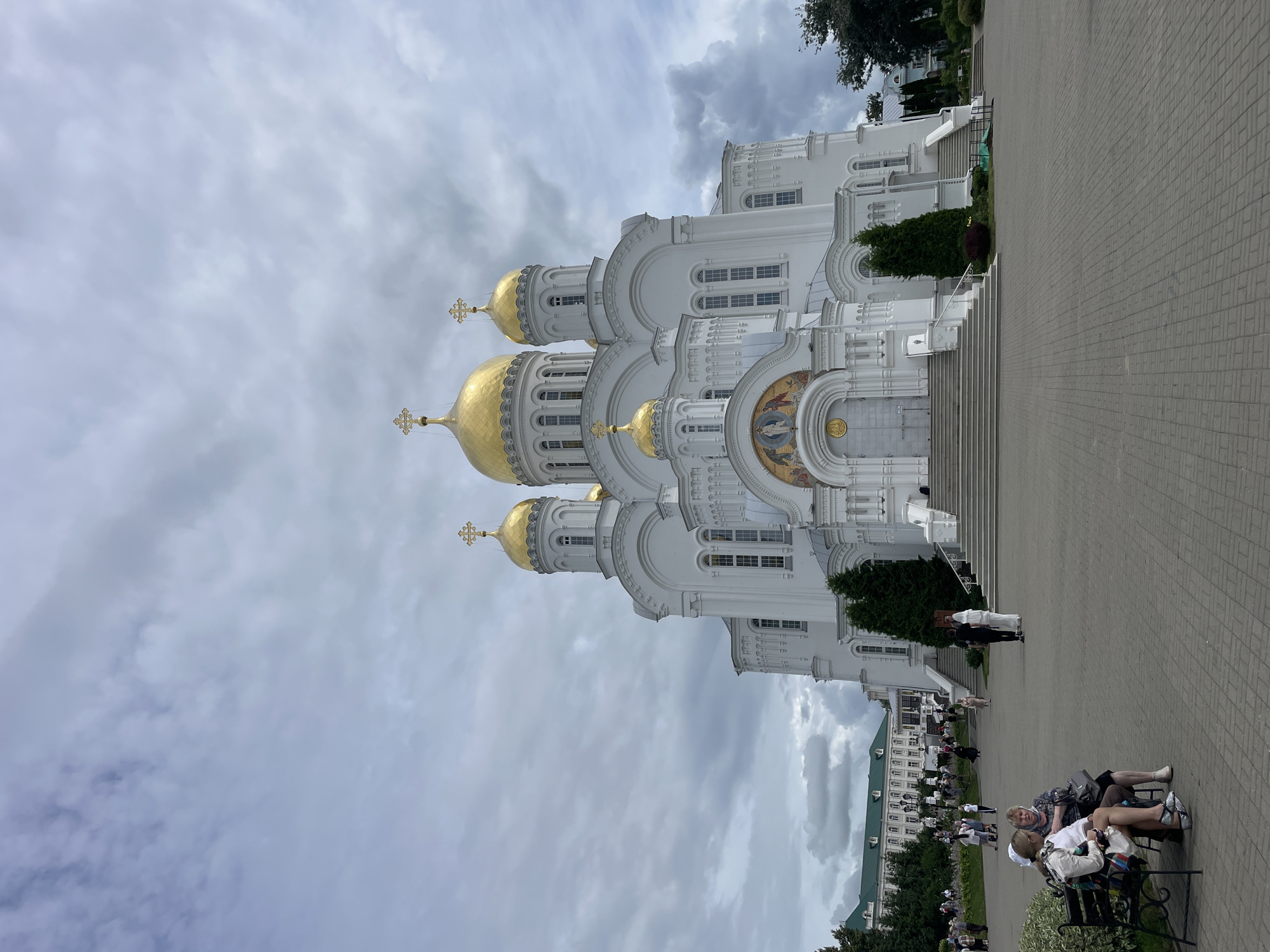 The image shows the Holy Trinity Cathedral in the city of Diveyevo, Russia, under a cloudy sky. The large, white cathedral features multiple golden domes topped with crosses. The entrance has a wide staircase leading up to ornate doors, with a mosaic above the entrance. Several people are visible in the foreground and background, including two women sitting on a bench on the left, and others walking around the plaza in front of the cathedral. To the left of the cathedral, there are other buildings with green roofs, and trees are scattered throughout the scene. The ground is paved with grey interlocking bricks.