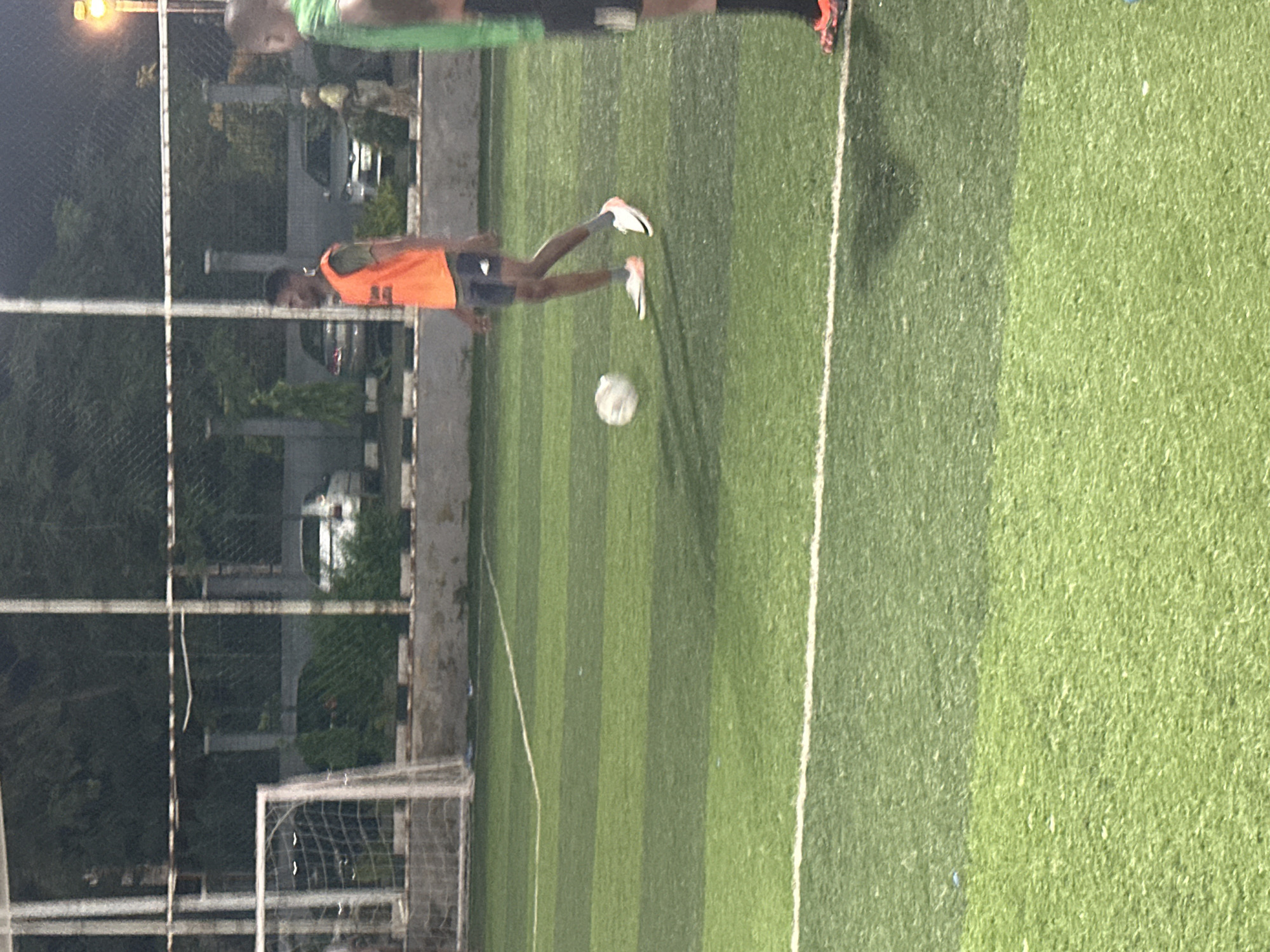 A low-angle, medium shot captures a football field in Abuja, Nigeria, at night. The artificial turf is illuminated by artificial lights, casting shadows across the green surface. In the mid-ground, a man wearing an orange training bib and dark shorts stands with a soccer ball at his feet, appearing to be in the process of kicking it. His athletic shoes are white with pink accents. To the right, another person, partially visible, wears a green top and black shorts, with orange and black cleats. In the background, a chain-link fence encloses the field, behind which trees and vehicles are dimly visible. A goalpost with a net is positioned to the left. The scene suggests a casual football training or match occurring after dark.