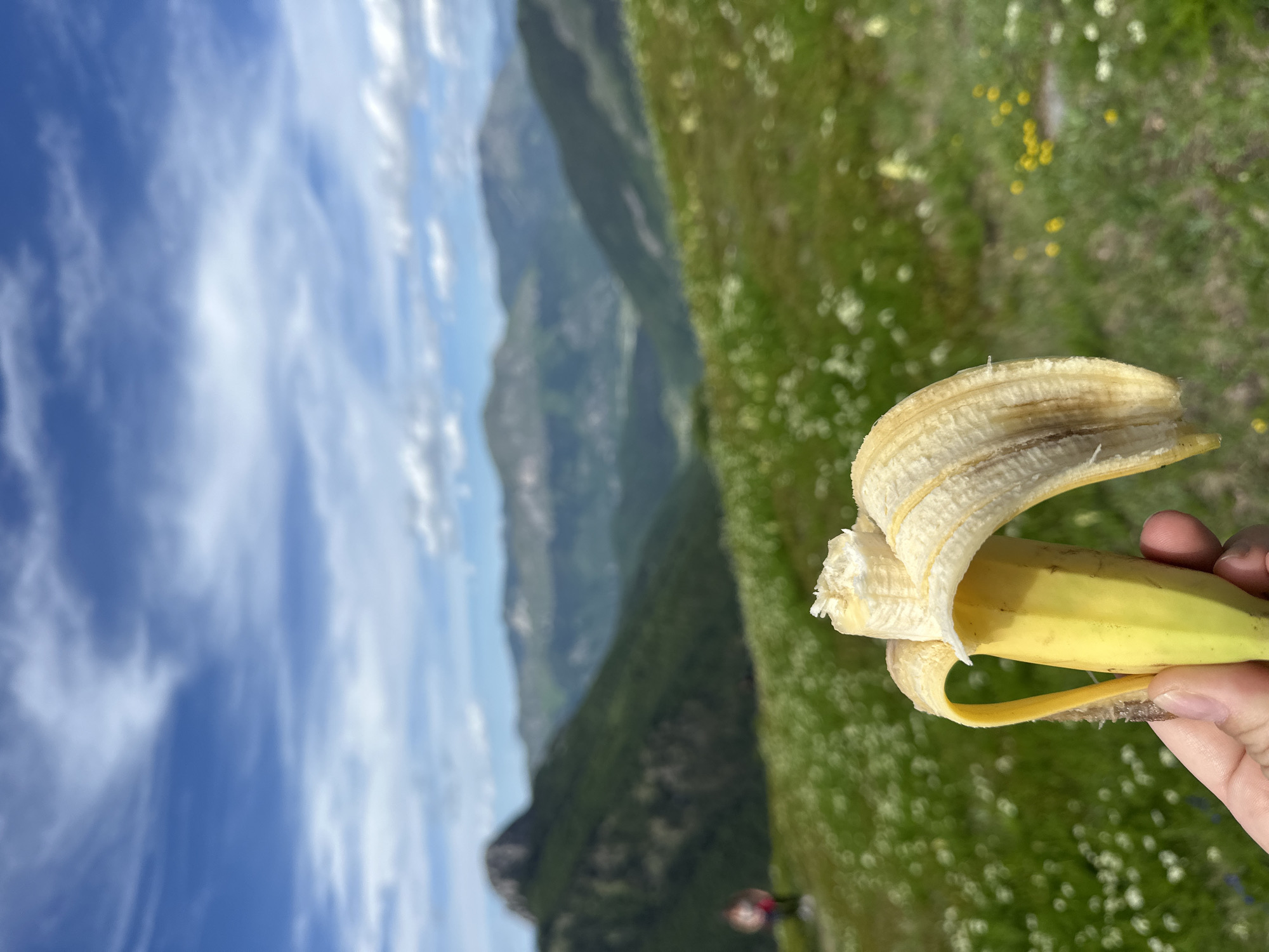 A partially peeled banana is held in a person's hand in the foreground. The banana is yellow with some brown spots. The background shows a lush green mountain landscape with scattered white wildflowers. In the far distance, a figure is visible on a hillside. The sky is blue with white, scattered clouds. The scene is set in the mountains near Dolja, Montenegro.