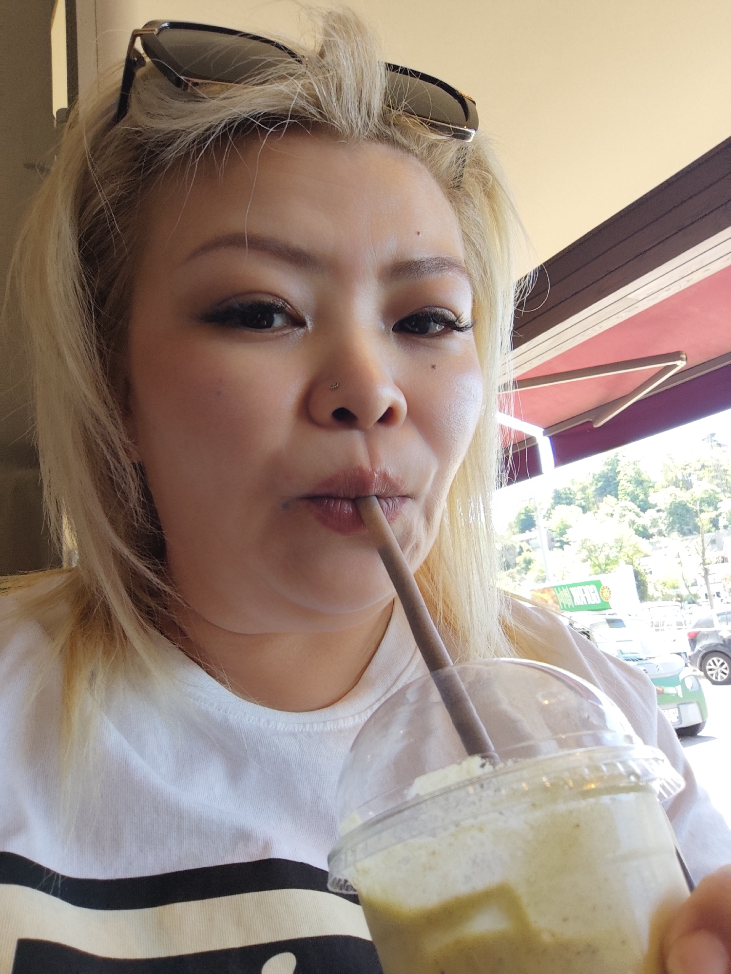 A woman with blonde hair, sunglasses on her head, and a nose piercing is drinking a beverage with a straw from a clear plastic cup. The cup contains a pale yellow liquid with some darker green sediment at the bottom. She is wearing a white t-shirt with a black and beige pattern.
The background shows a street scene with cars and green trees. A partial awning with a red and dark wood design is visible above the woman. A sign with green and white text is also in view. The location is in Sarıyer, Turkey.