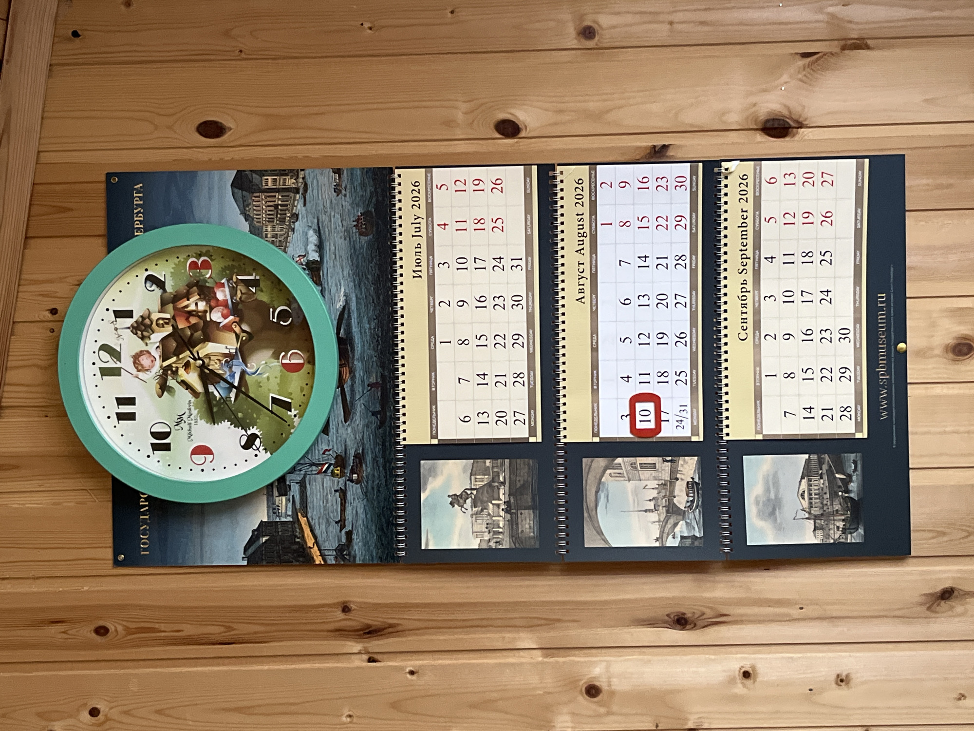 A round clock with a turquoise frame is mounted on a wooden wall. The clock face features Roman numerals and an image of cherubs and buildings. Below the clock, three calendars are displayed, each with a photograph and a monthly schedule for 2026. The top calendar shows July, the middle one shows August, and the bottom one shows September. The August calendar has the number 10 circled in red. The overall scene suggests a domestic or office setting decorated with historical and time-keeping elements. There is no indication of the city of Ryabovo, Russia.