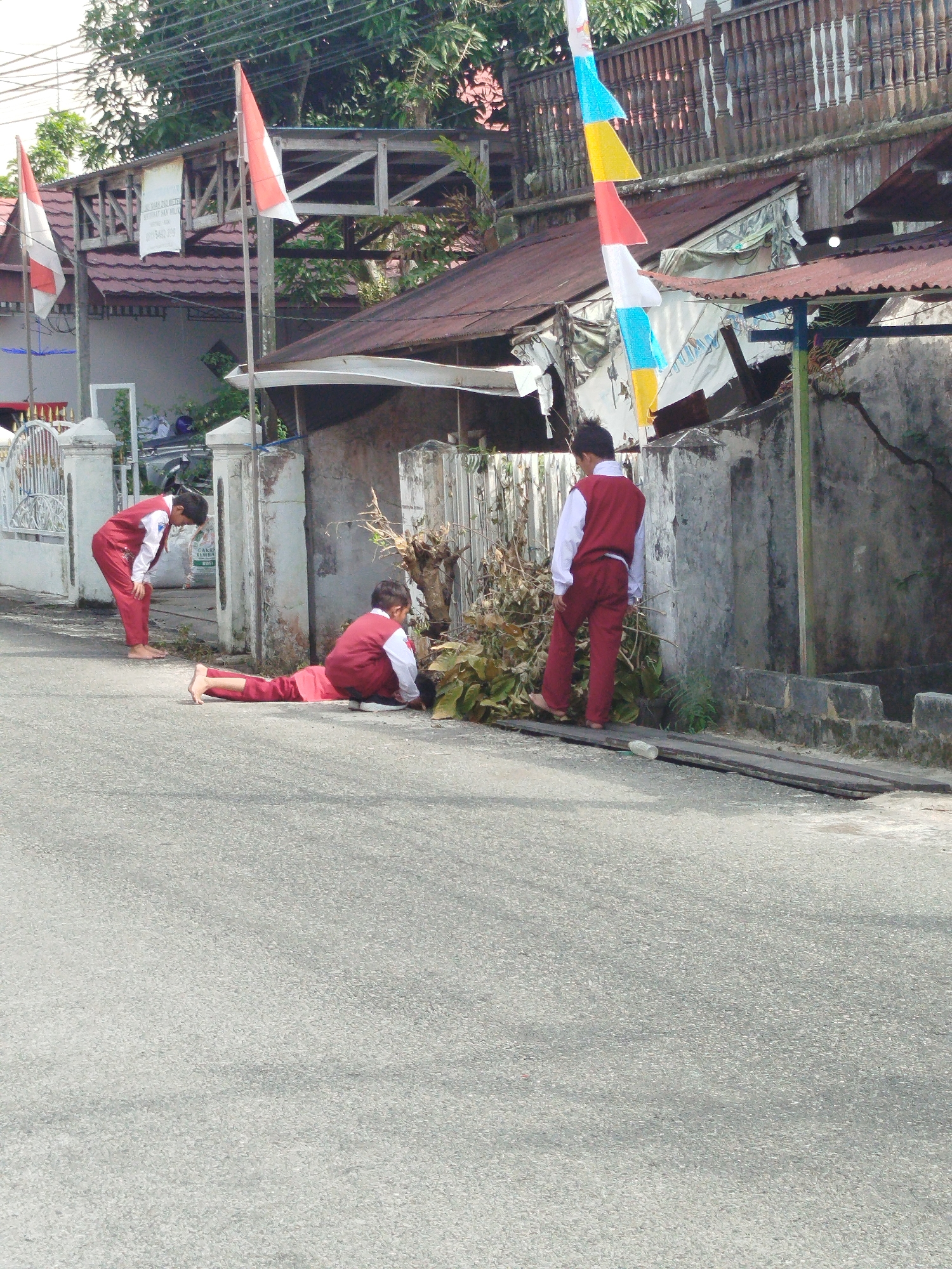 A boy in a red and white uniform lies on his stomach on the asphalt road in front of a concrete wall and a wooden fence. He is near some dried vegetation and sticks. His legs are extended straight out behind him and he is looking down. Another boy in a similar uniform stands behind him, facing away from the camera, and appears to be looking at the dried vegetation. To the left, further down the road, a third boy in a red and white uniform is bent over and looking towards the ground. Red and white flags are visible in the background. The scene appears to be a street in Balikpapan, Indonesia, possibly near a school or residential area, given the uniforms and the architecture of the buildings.