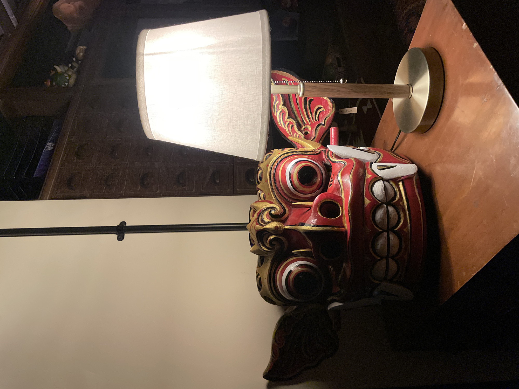 An indoor scene showcases a vibrant, intricately carved Balinese-style mask positioned next to a lit table lamp on a wooden surface. The mask, likely depicting a mythological creature such as Barong or Rangda, is predominantly red with elaborate gold, black, and white accents, including swirling patterns and large, bulging eyes with concentric circles. Its fierce expression features prominent white fangs on one side.
The lamp has a simple, cylindrical white fabric shade, a light wooden stem, and a round, brass-colored metal base. A small pull chain is visible near the top of the shade, suggesting it's an on/off mechanism. The lamp casts a bright glow, illuminating the mask and the immediate area of the wooden table or shelf it rests upon.
In the darker background, to the left of the mask and behind the lamp, dark wooden furniture, possibly shelves or a cabinet, is visible. Faint shapes of other objects, perhaps decorative figurines or plush toys, can be discerned on these shelves. To the far left, a plain, light-colored wall features a horizontal black rod with a clip attached. On the wooden surface near the lamp's base, a small, partially visible label or sticker can be seen with what looks like "5mil". The overall lighting suggests an evening or nighttime setting indoors, with the lamp as the primary light source. The style of the mask aligns with the provided location context of Mengwi, Indonesia, known for its rich cultural traditions and artistry.