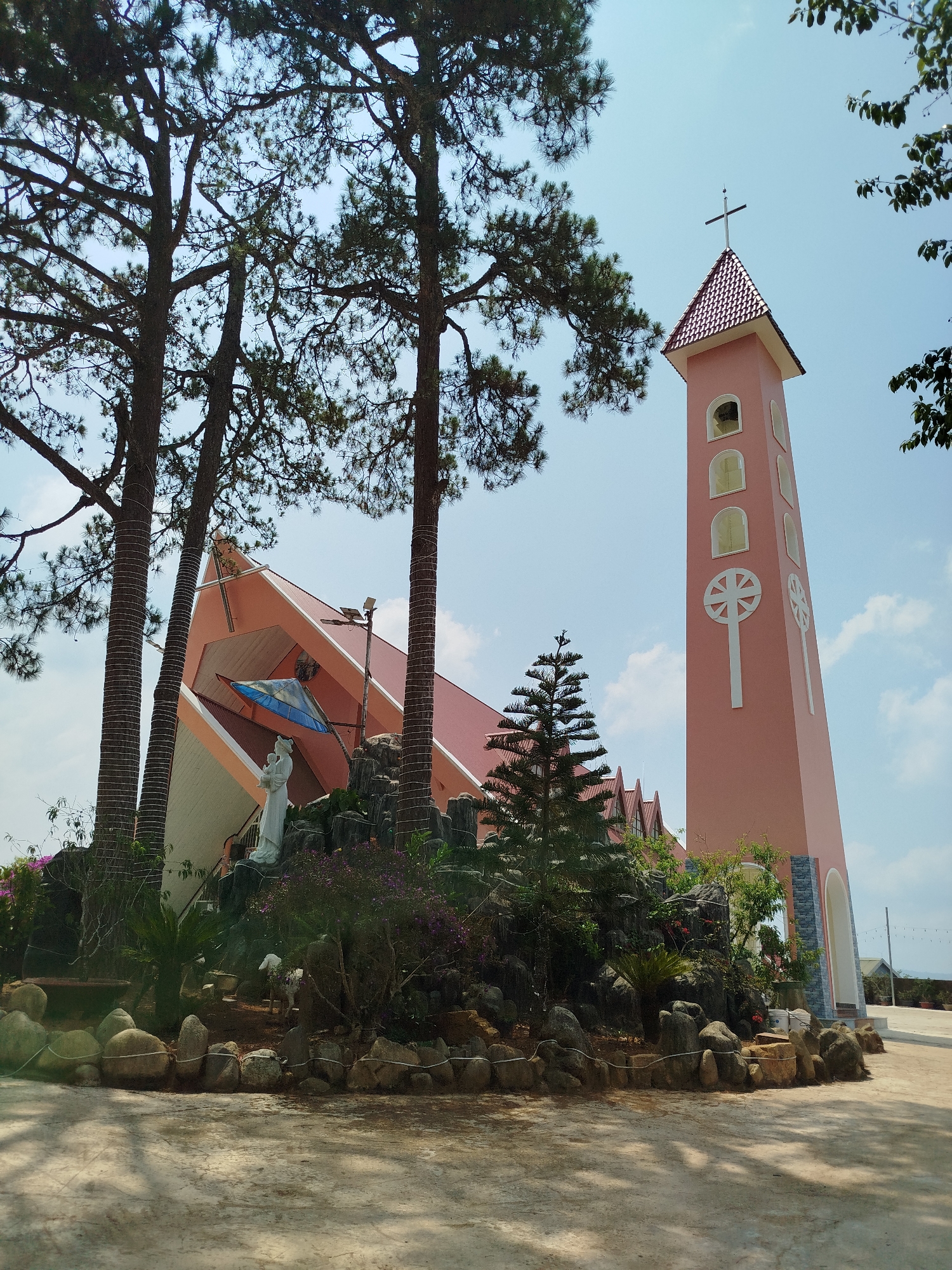 The image depicts a pink church with a tall bell tower in Đa Thọ, Vietnam. The church has a modern architectural design with sharp angles and a light pink exterior. A bell tower stands adjacent to the church, featuring a cross at its apex and white decorative elements. The church is surrounded by lush greenery, including tall trees, flowering plants, and decorative rocks. A white statue of the Virgin Mary holding the baby Jesus is positioned in front of the church. The ground in front of the church is paved. The sky is blue with scattered clouds.