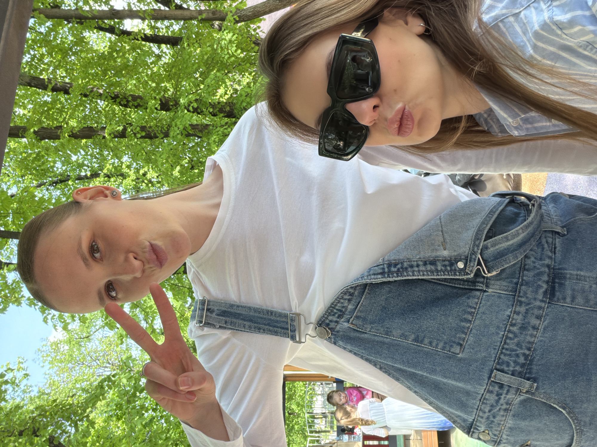 Two young women are posing for a selfie. One woman is wearing a white long-sleeved shirt and denim overalls and is making a peace sign. The other woman is wearing sunglasses and a blue and white striped shirt and is pouting her lips. They appear to be outdoors, in a park or recreational area, with trees visible in the background. There is also a playground visible in the background. The picture may have been taken in Moscow, Russia.
