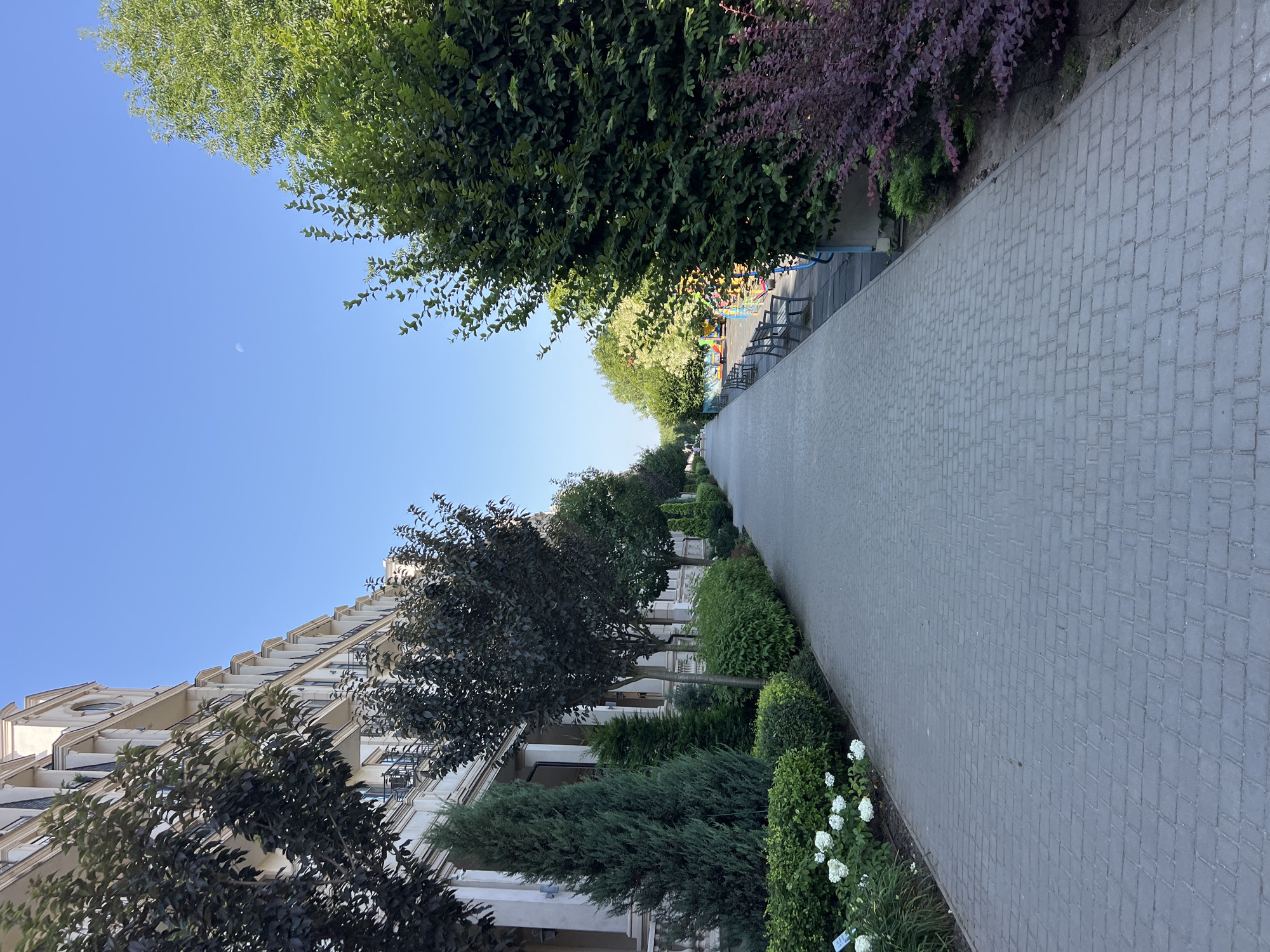The image depicts a pedestrian walkway in Fontanka, Ukraine, adjacent to a multi-story building with arched balconies. The walkway is paved with gray rectangular bricks and is lined with various trees and shrubs. On the left side, there is a row of manicured evergreen trees and bushes, with white flowers blooming near the base. Further down the walkway, a playground with colorful equipment is visible on the right. Several park benches are placed along the walkway. The sky is clear and blue, with a faint crescent moon visible. The scene appears to be in the daytime, with ample natural light.