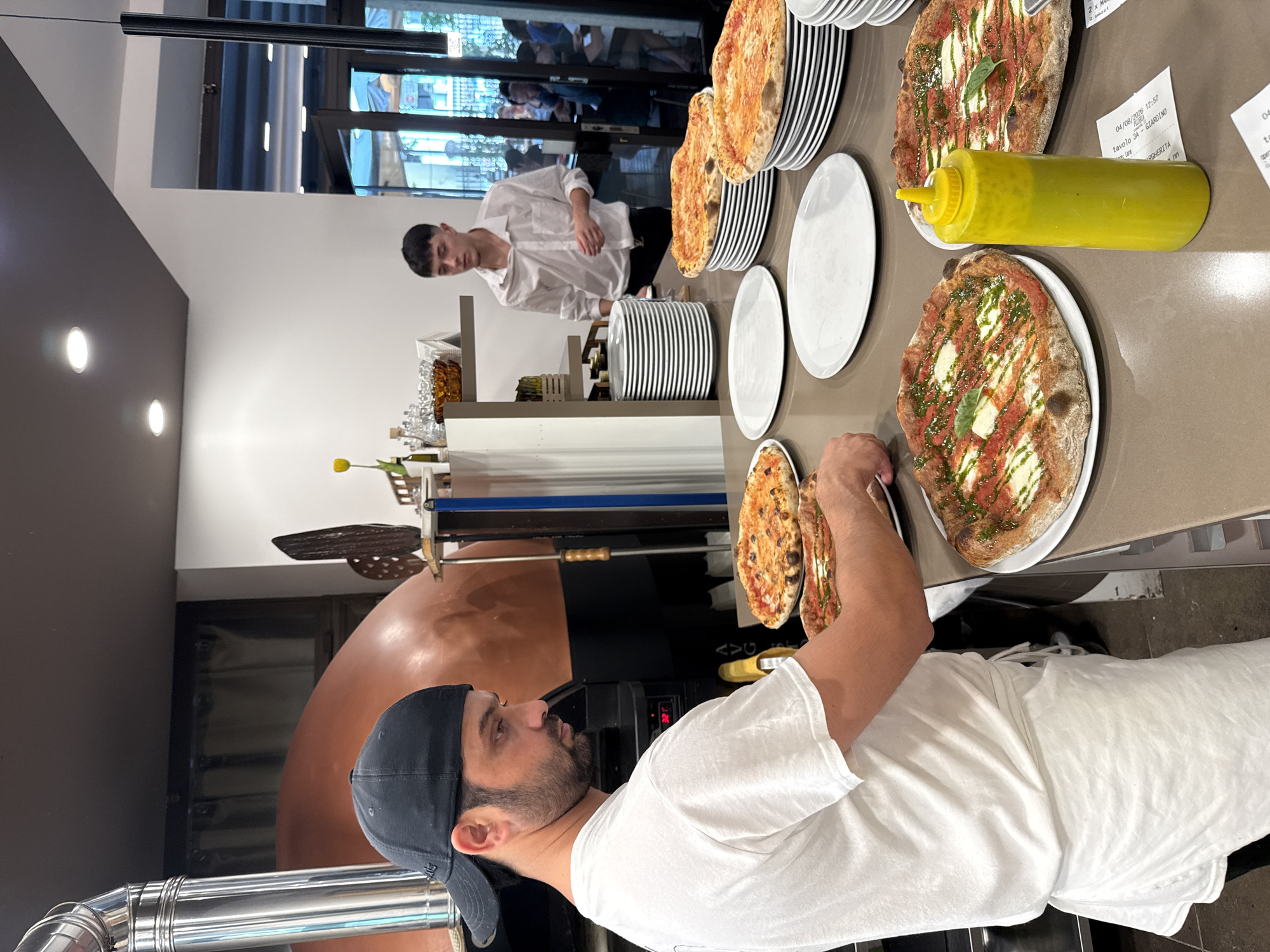 The image depicts an indoor scene, likely a restaurant kitchen or food preparation area, with a focus on pizza. In the foreground, a man wearing a white t-shirt and a black baseball cap is seen from the chest up, facing to his right and looking down at pizzas on the counter. He appears to be a pizza maker or chef. Several pizzas are visible on white plates and stacked on black platters. To the right of the man, a bright yellow squeeze bottle, possibly containing a sauce or condiment, stands upright. In the background, another young man in a white collared shirt stands behind a stack of white plates, looking towards the front. Behind him, a large, dome-shaped pizza oven, possibly made of copper, is partially visible. The environment appears to be clean and functional, with various kitchen items and stacks of plates suggesting active food service. There are glimpses of outdoor scenery through windows in the far background, hinting at a city environment, potentially Trento, Italy, as indicated by the presence of receipts with Italian text and possible local establishment clues.