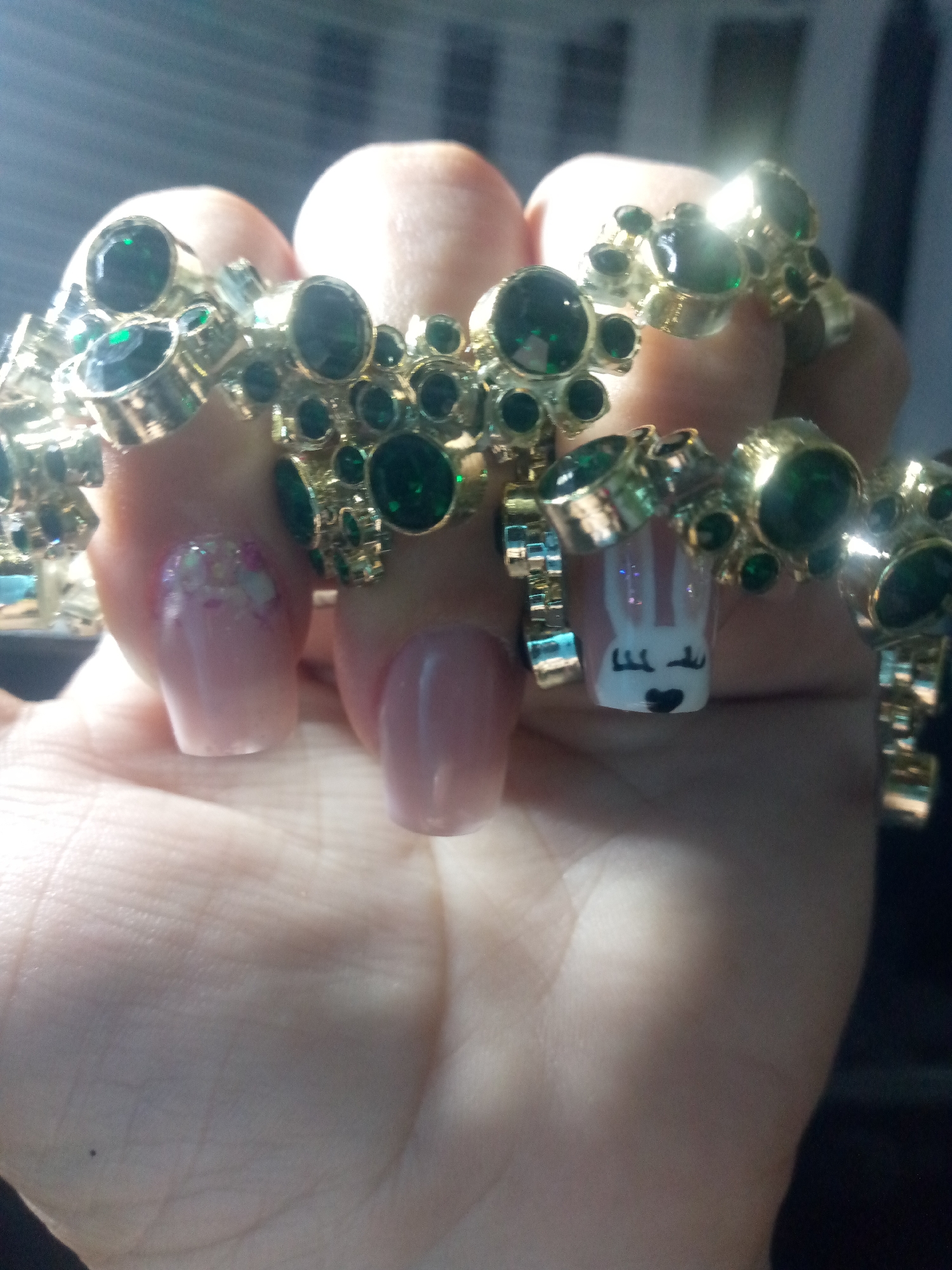 A close-up shot shows a person's hand adorned with a gold-colored bracelet featuring green stones. The hand has manicured nails. One nail is decorated with a white bunny face, complete with ears, eyes, and a heart-shaped nose. Another nail is adorned with a floral pattern. The background is blurred, but appears to be indoors, possibly near a window. No specific location within any city can be determined from this image.