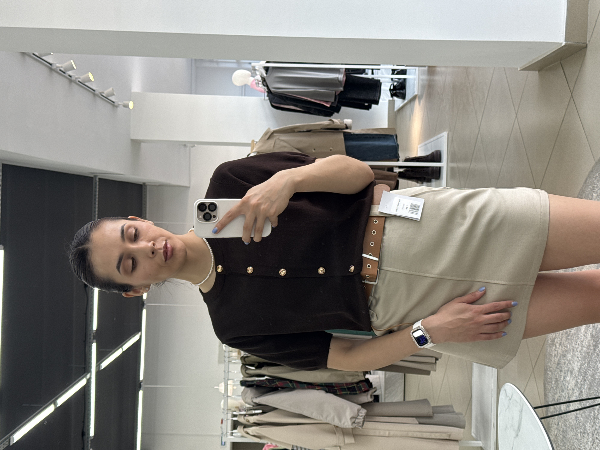 A woman is taking a selfie in what appears to be a clothing store. She is wearing a brown short-sleeved cardigan with gold buttons, a light tan mini skirt with a brown belt, and a pearl necklace. She is holding a white smartphone. Racks of clothing are visible in the background, along with various display racks. The location is likely a clothing store in a shopping center, but there's no definitive information on the city or the specifics of the location within Votkinsk, Russia from the image provided.