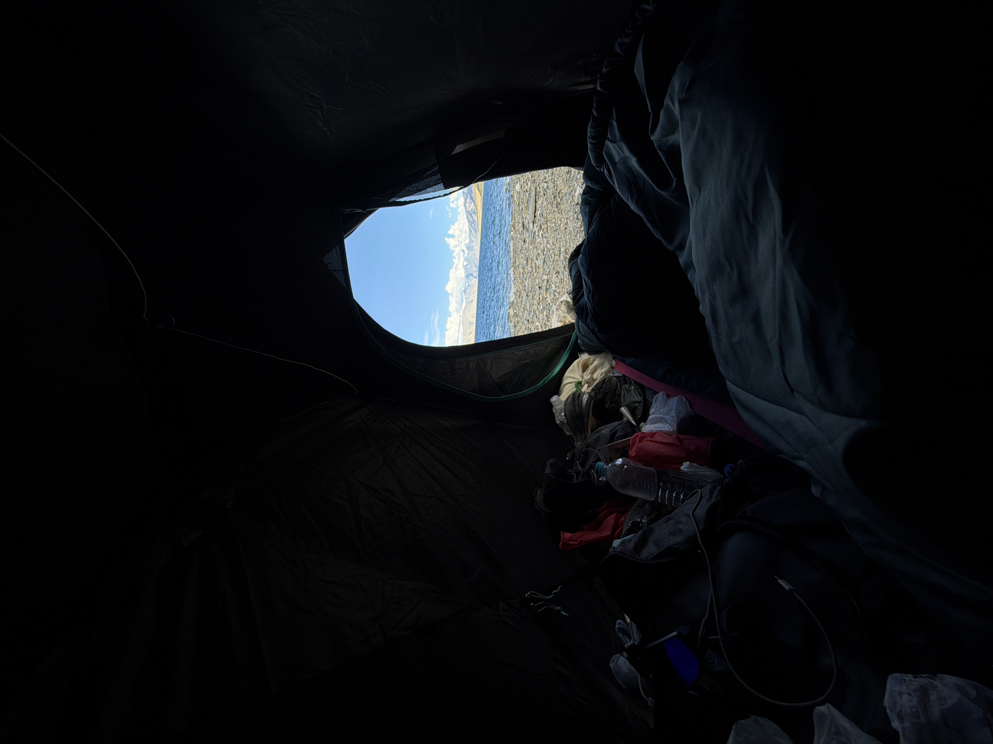 The image is taken from inside a dark tent, looking out through the open entrance. The foreground is filled with camping gear, including sleeping bags, bags, and a water bottle. The entrance of the tent frames a scenic view of a lake with snow-capped mountains in the distance. The sky is blue with scattered clouds. The terrain outside the tent appears to be rocky. The overall impression is one of a remote, natural landscape.