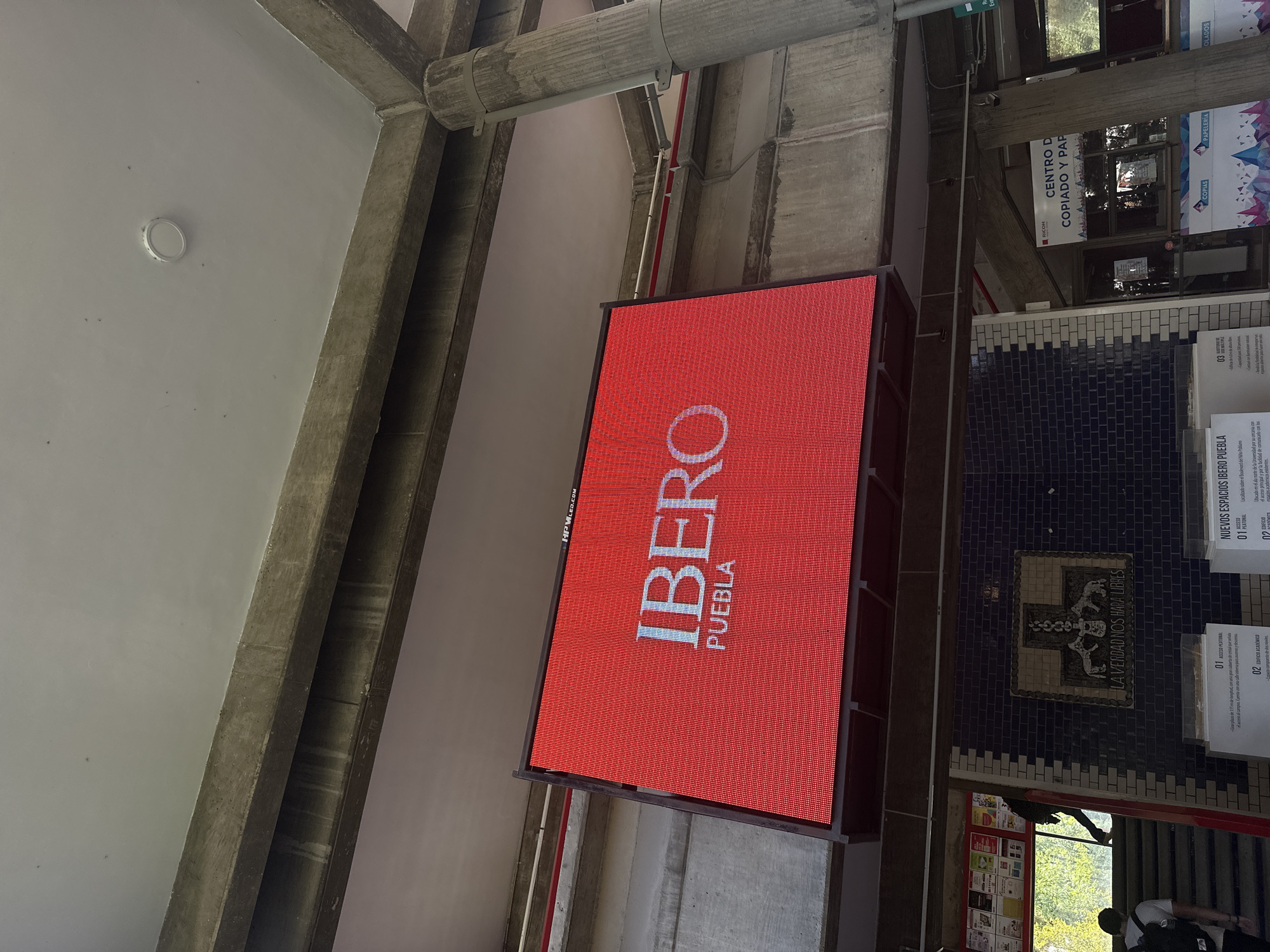This image captures an indoor architectural space, likely within a university or public building, characterized by exposed concrete beams and a light-colored ceiling. A prominent feature is a large, vertical red LED screen displaying the white text "IBERO PUEBLA" in bold letters, with "HPMLED.COM" visible in small text at the top left.
To the right of the screen, a wall is decorated with dark blue and white tiles, featuring a mosaic design that includes the motto "LA VERDAD NOS HARA LIBRES" (The truth will set us free). Several informational signs are posted on this wall. One sign advertises a "CENTRO DE COPIADO Y PAPELERÍA" (Copying and Stationery Center) and mentions "RICOH," "COPIAS," "PAPELERÍA," and "ENMICADOS" (laminations). Another sign details "NUEVOS ESPACIOS IBERO PUEBLA" (New Ibero Puebla Spaces), with bullet points for "01 ACCESO PEATONAL" (Pedestrian Access) and "02 EDIFICIO ACADÉMICO" (Academic Building), along with descriptive text for each.
At the bottom right of the image, parts of two people are visible, suggesting activity and movement in the area. The setting appears to be a modern and functional environment, consistent with a university campus like Iberoamericana University Puebla in San Bernardino Tlaxcalancingo, Mexico, as indicated by the prominent signage. The lighting suggests it is daytime.