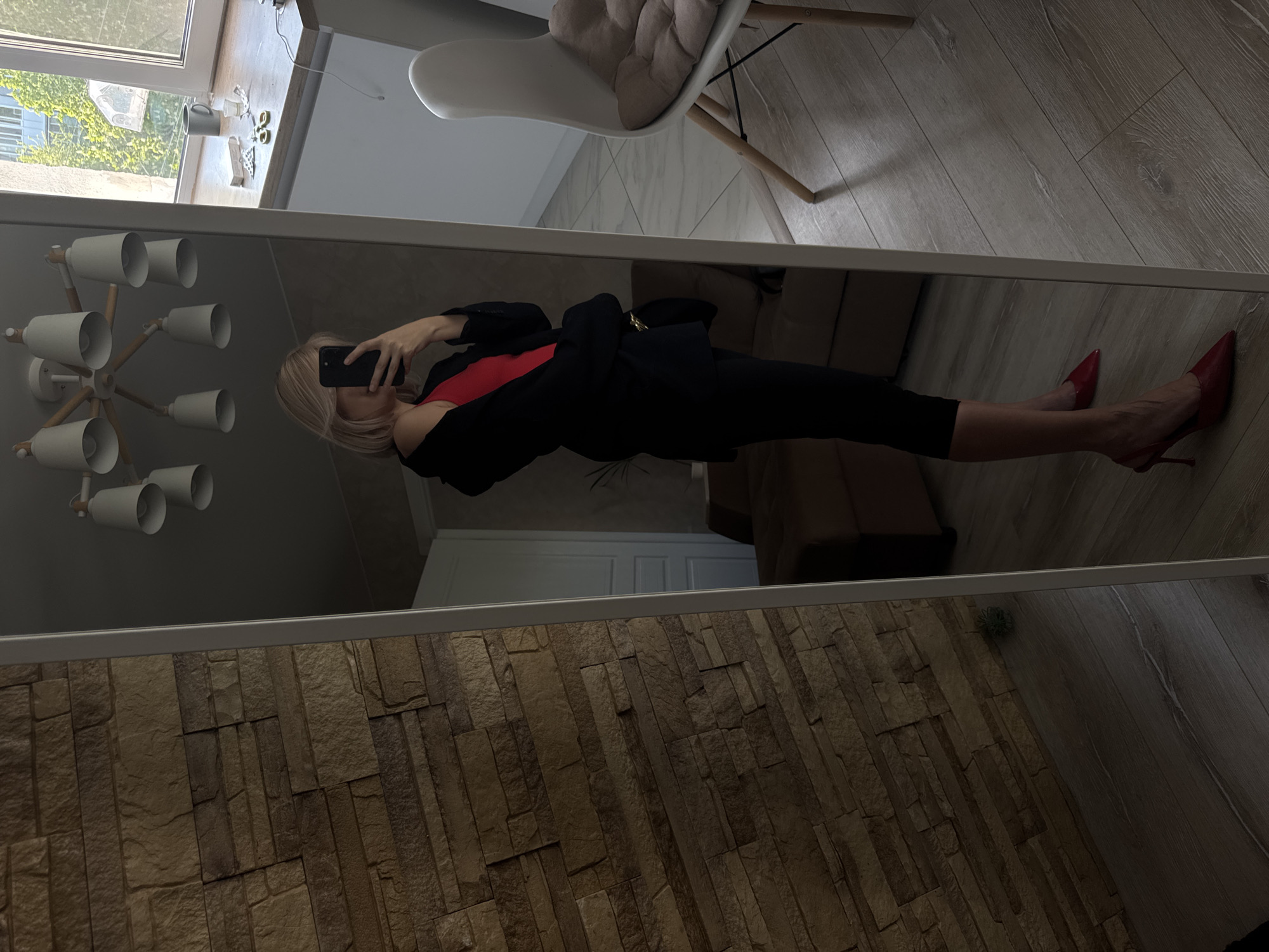 A woman is reflected in a mirror, taking a photograph of herself with a mobile phone. She has blonde hair and is wearing a black blazer over a red top, with black leggings or trousers. She is also wearing red high heels. The mirror's reflection shows parts of a room, including a white chair, a ceiling light fixture, and a textured wall. The location is not discernible from the image.
