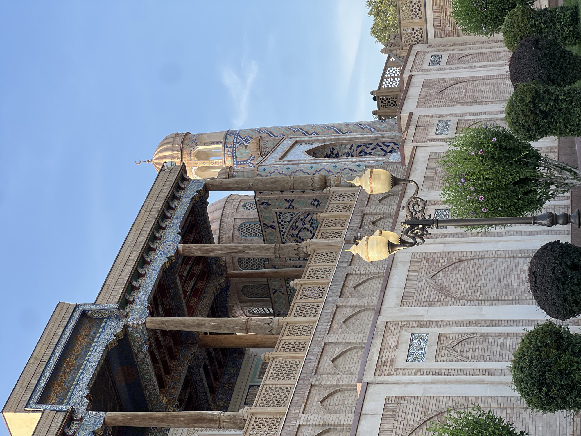 The image captures an architectural detail of a building, likely a mosque or madrasah, in Samarkand, Uzbekistan. The perspective is from below, looking up at the building's facade and a minaret against a clear blue sky. The architecture is ornate, featuring intricate tilework in shades of blue, white, and turquoise, forming geometric patterns and arabesques.
The main subject is the building itself, with its brickwork and decorative elements. A tall, slender minaret, adorned with similar colorful tiles, rises prominently. Below the minaret, a domed structure is partially visible. The building's facade is constructed of light-colored bricks, with arched decorative niches and bands of tilework. Several wooden columns support an ornate overhang. A decorative, openwork lattice structure, possibly a balcony or part of a staircase, is visible.
Decorative lamps with yellowish glass shades are mounted on a lamppost, casting a warm glow, suggesting it might be late afternoon or early evening, or that the lamps are illuminated even during the day for aesthetic purposes. The surrounding area is landscaped with well-maintained green bushes and trees, adding to the serene and cultivated atmosphere. The sky is a bright, clear blue with a few wispy clouds, indicating pleasant weather.
There is no visible text in the image. The overall impression is one of historical grandeur and intricate craftsmanship, characteristic of Central Asian Islamic architecture. The scene appears calm and still, with no people or explicit activity depicted, focusing solely on the architectural beauty of the site.