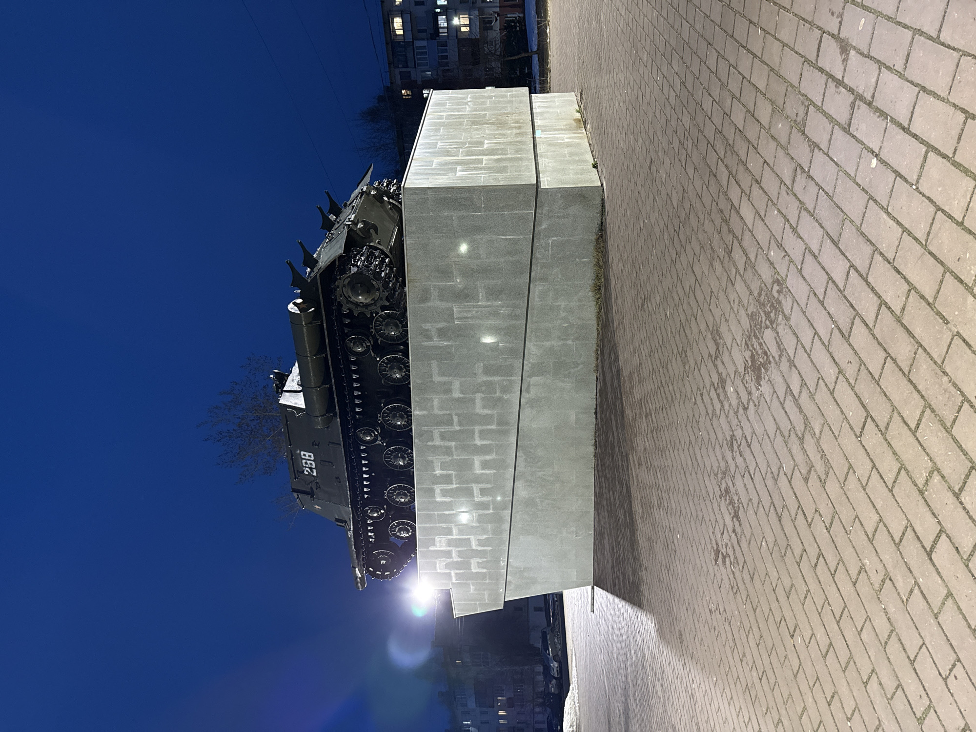 The image shows a T-34 tank mounted on a gray brick pedestal. The tank is oriented with its front facing the viewer's left. The tank has the number "288" painted on its side. The sky behind the tank is a deep blue. To the right of the pedestal is a paved walkway. In the background are buildings with lighted windows. This memorial is located in the city of Chaikovsky, Russia. There are no people in the image.