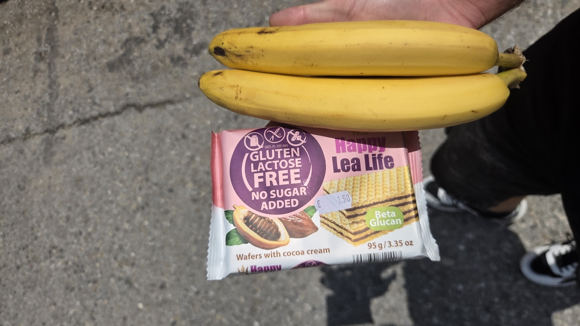 A pair of yellow bananas are held in a person's right hand. The bananas are positioned vertically, with their stems pointing downwards. To the left of the bananas is a package of wafers. The package is mostly pink and white with text and images of cocoa pods and wafers. The text on the package reads "Happy Lea Life", "Gluten Free", "Lactose Free", and "No Sugar Added". Below that, it says "Wafers with cocoa cream" and "95g/3.35 oz". A price tag of "€ 1.50" is visible on the package. The background appears to be a grey asphalt surface. The person's legs and feet are visible at the bottom of the image, wearing black pants and white sneakers.