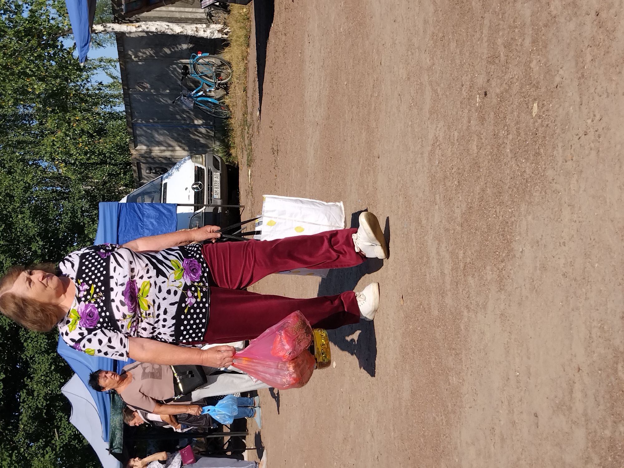 A woman walks outdoors in the city of Irshansk, Ukraine. She is wearing a white shirt with black polka dots and floral patterns, and burgundy pants. She carries a pink plastic bag filled with items and a yellow container. In the background, tents and a white van are visible. A blue bicycle is parked next to a fence. The ground is covered in dirt and gravel.