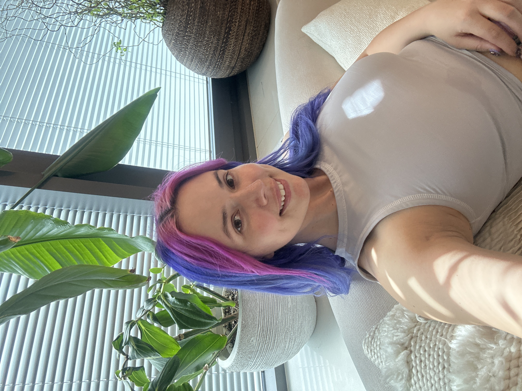 A woman with blue and pink hair lies on a beige couch next to a large window with blinds. The location is in a building in the City of Zagreb, Croatia. A large potted plant with green leaves is visible next to the window.